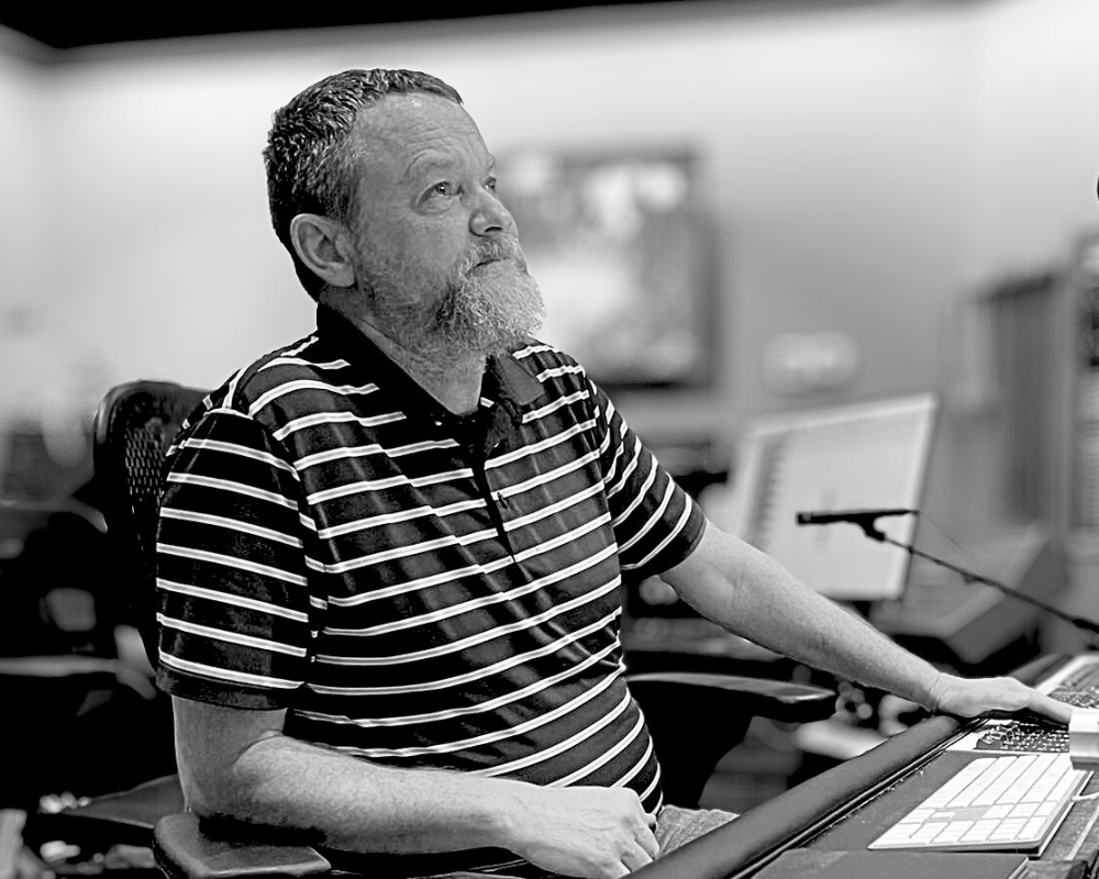 A bearded man in a striped polo shirt working at a music mixing console in a recording studio.