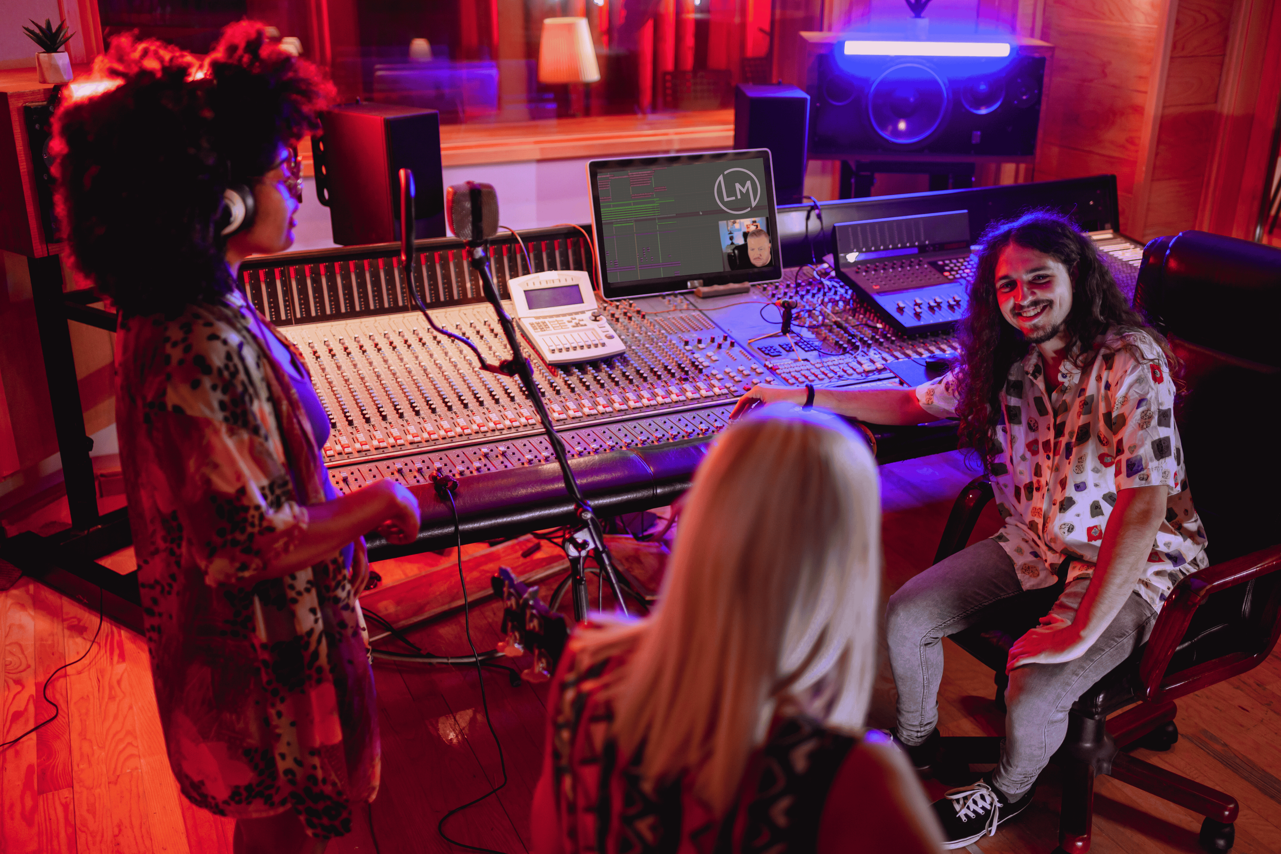 Three people in a recording studio with a large mixing console, computer screens, and professional audio equipment. One person with curly hair and leopard print shirt is standing and wearing headphones. Another person with long blonde hair is sitting with her back to the camera. The third person with long dark hair and a patterned shirt is sitting and smiling at the camera.