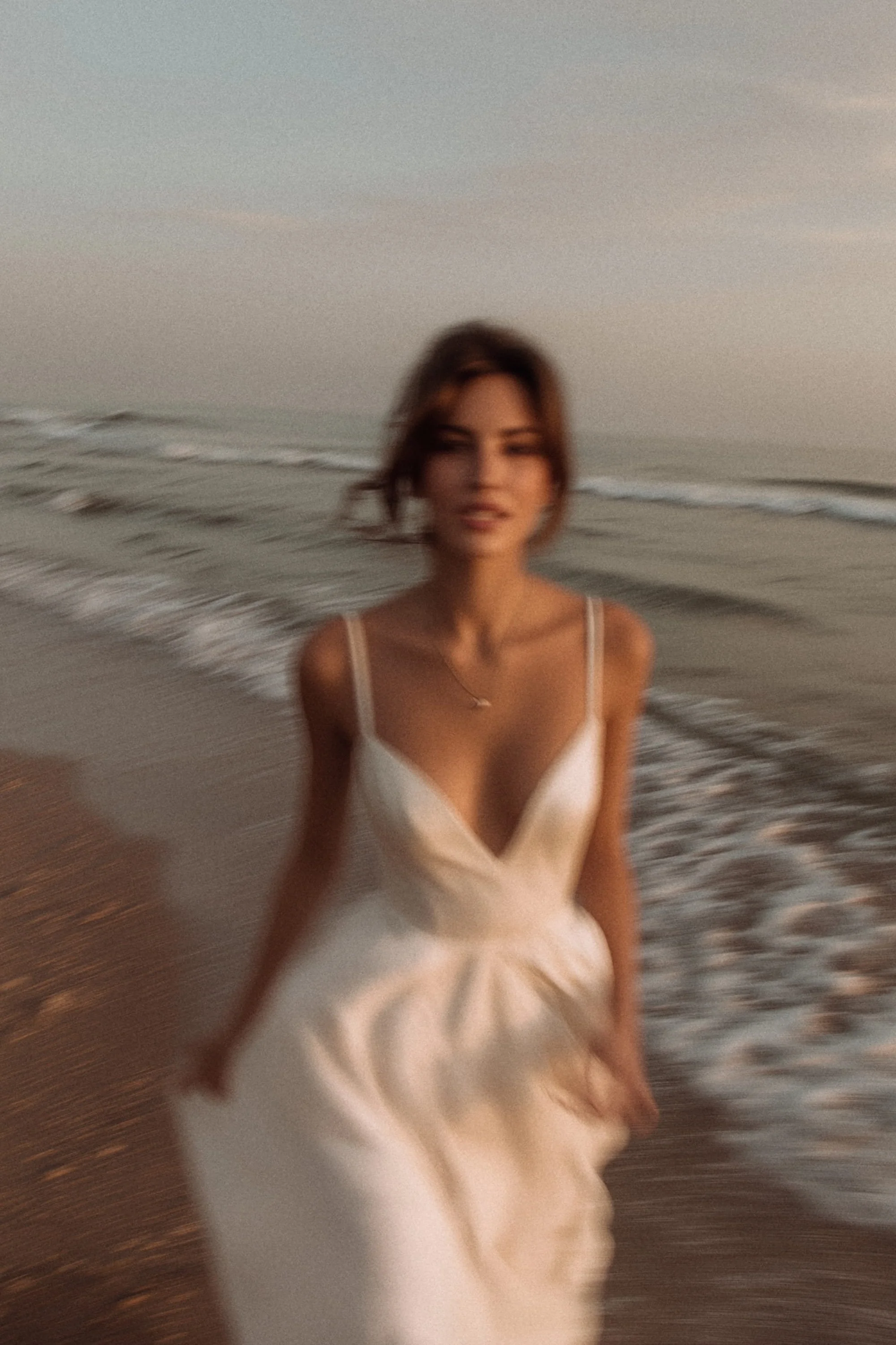 A woman in a white dress standing on a beach with waves in the background, during sunset.