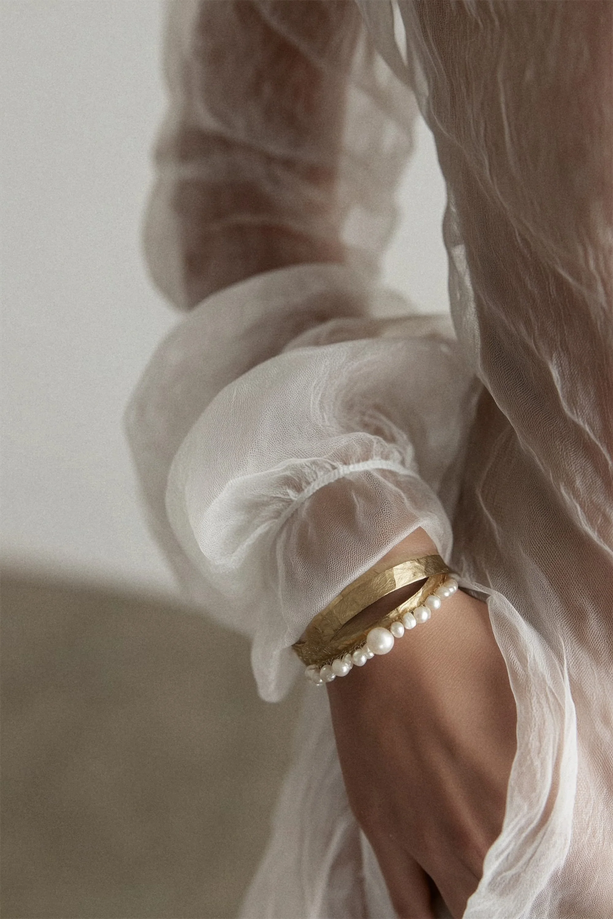 Close-up of a wrist adorned with a gold bracelet and a string of pearls, wearing a sheer, ruffled white blouse.