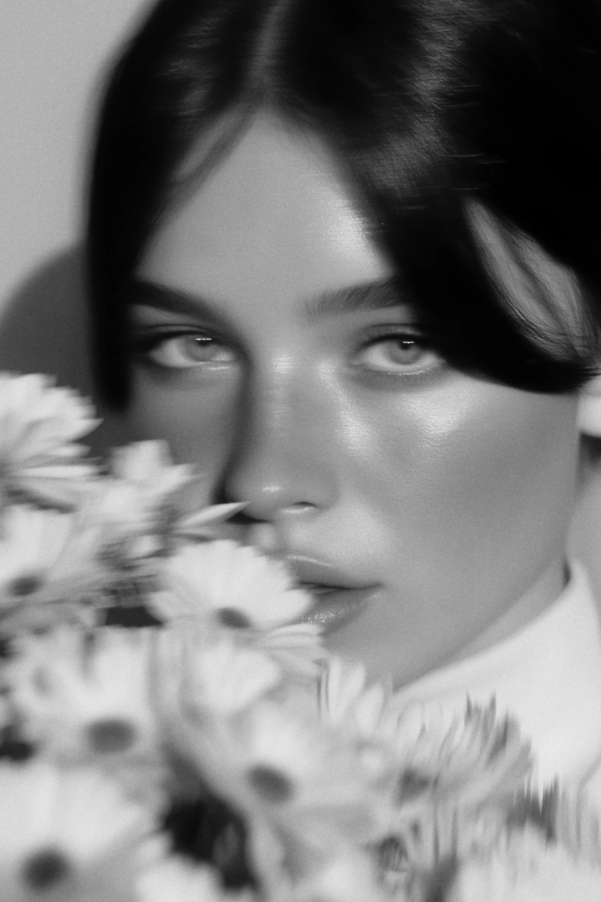 A black and white photo of a woman with short dark hair and striking features, partially obscured by flowers in the foreground.