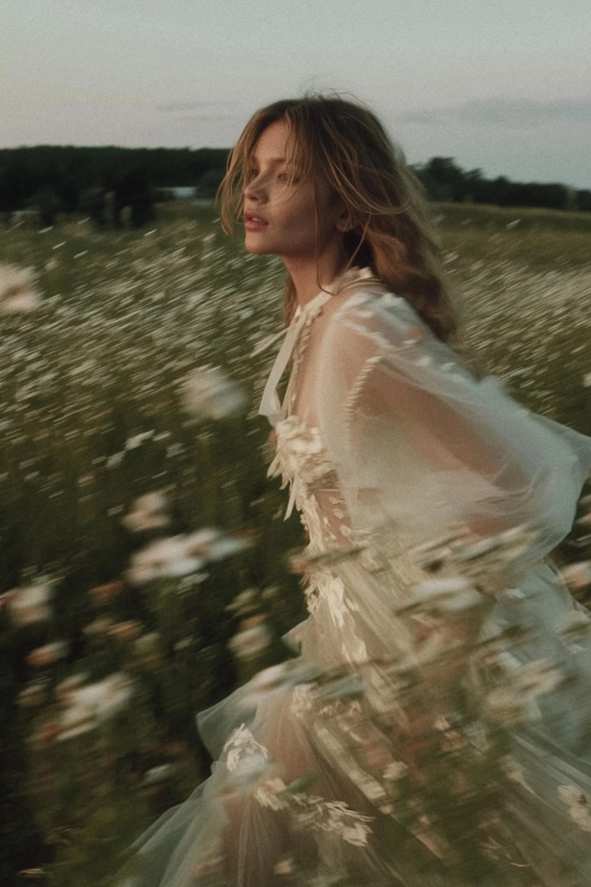 A woman with light brown hair running through a field of white flowers, wearing a flowing, sheer, light-colored dress during dusk.