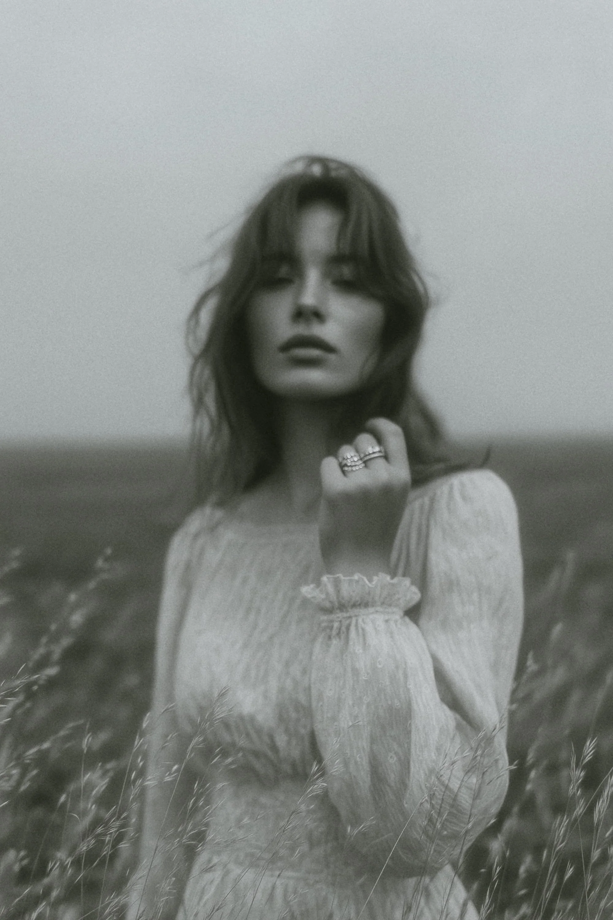 A woman with wavy hair standing in a field of tall grass, wearing a light-colored, long-sleeved blouse with ruffled cuffs, and holding her hand near her face with rings on her fingers.