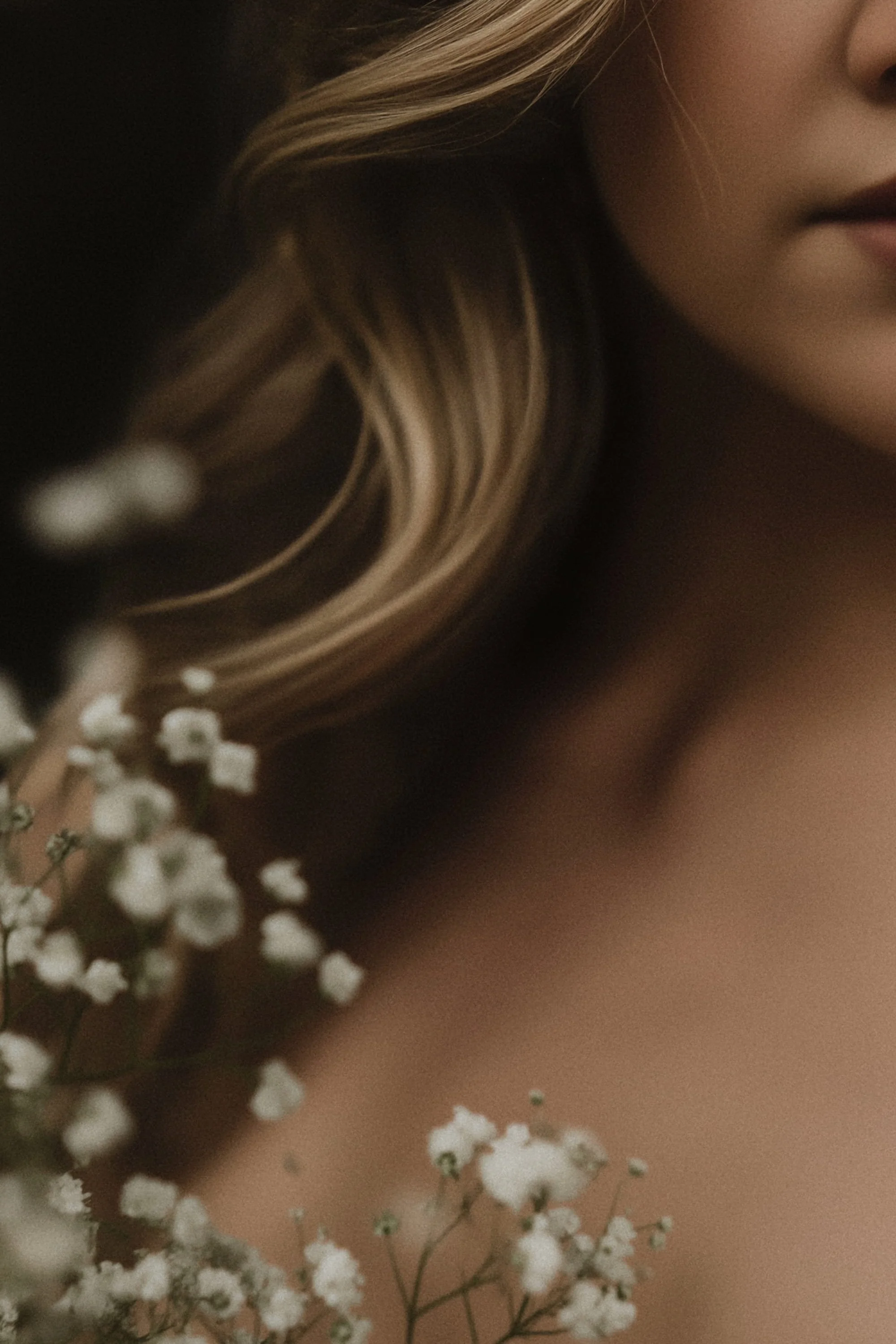 Close-up of a woman’s face and shoulder, with a focus on her blonde hair and part of her lips, next to white flowers.