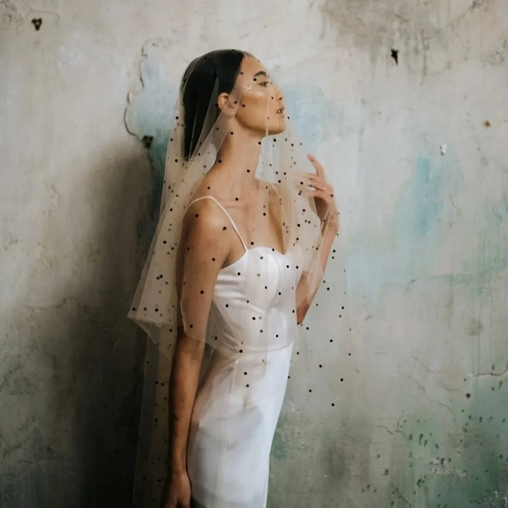 A woman in a white dress with a tulle veil decorated with black dots, posing against a textured wall.