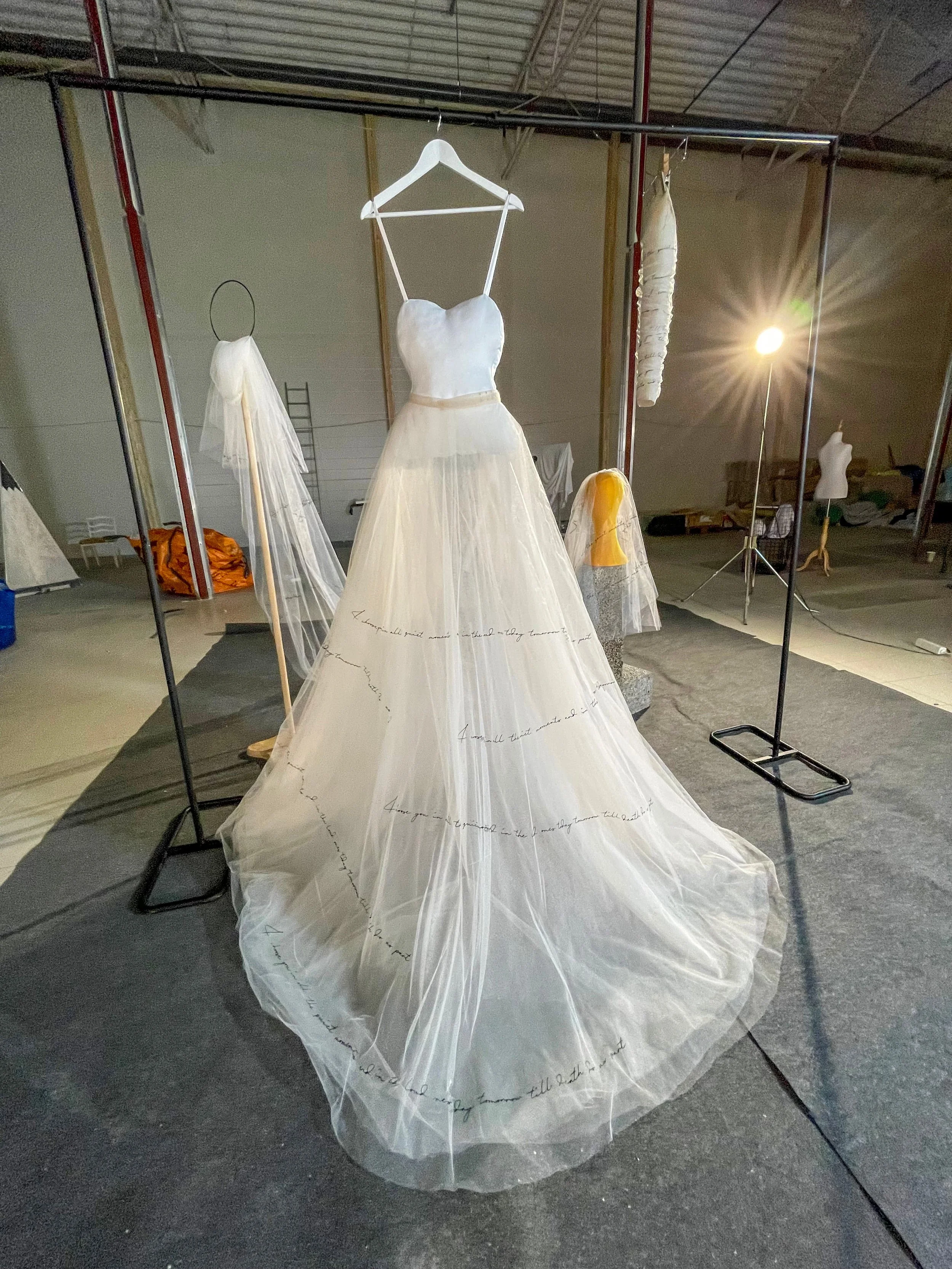 Design dress on a mannequin with a sheer, white fabric and handwritten text patterns, displayed in a studio setting with other dress forms and equipment.