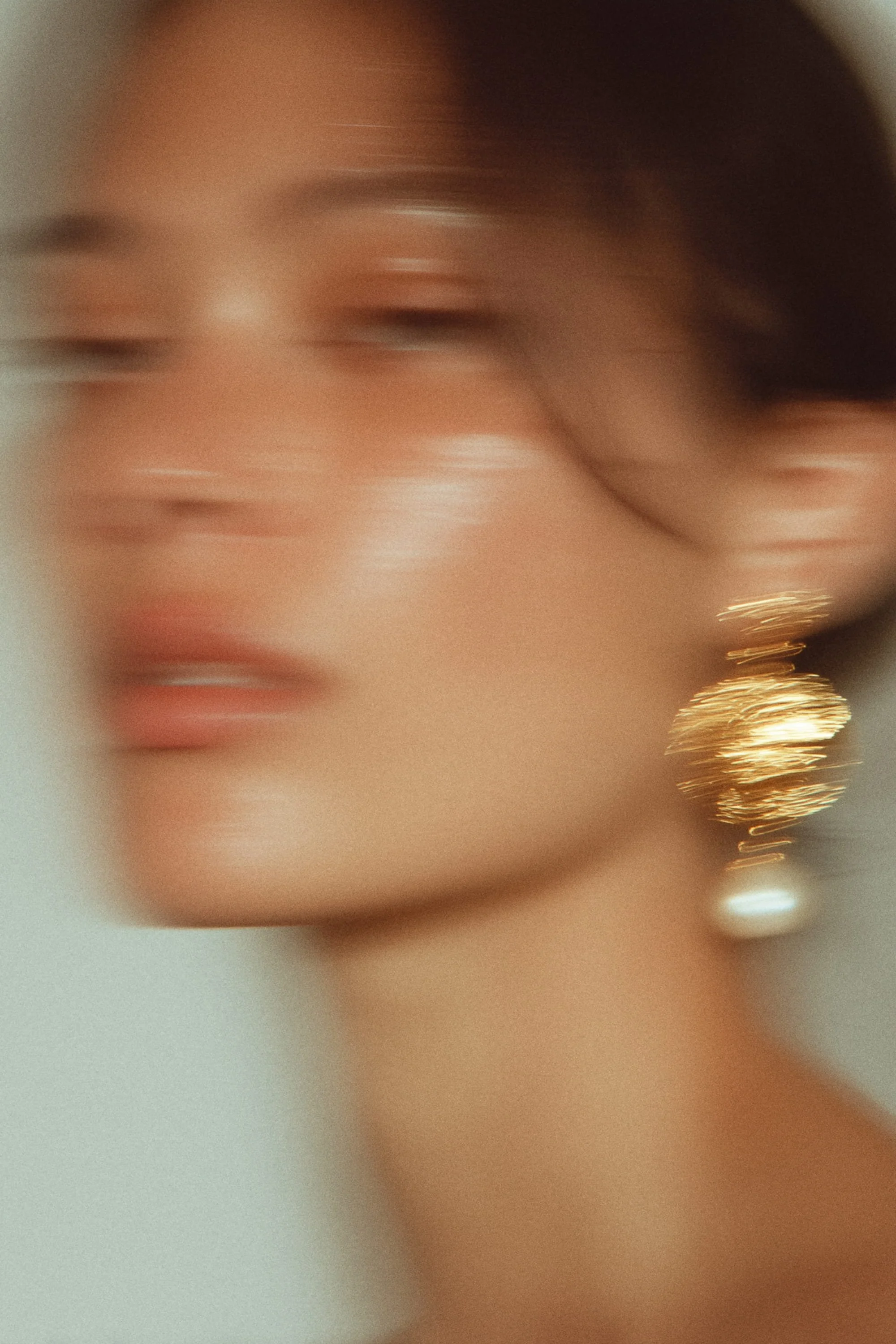 A blurred close-up portrait of a woman wearing a large gold earring with rhinestones and a pearl, with makeup and styled hair.