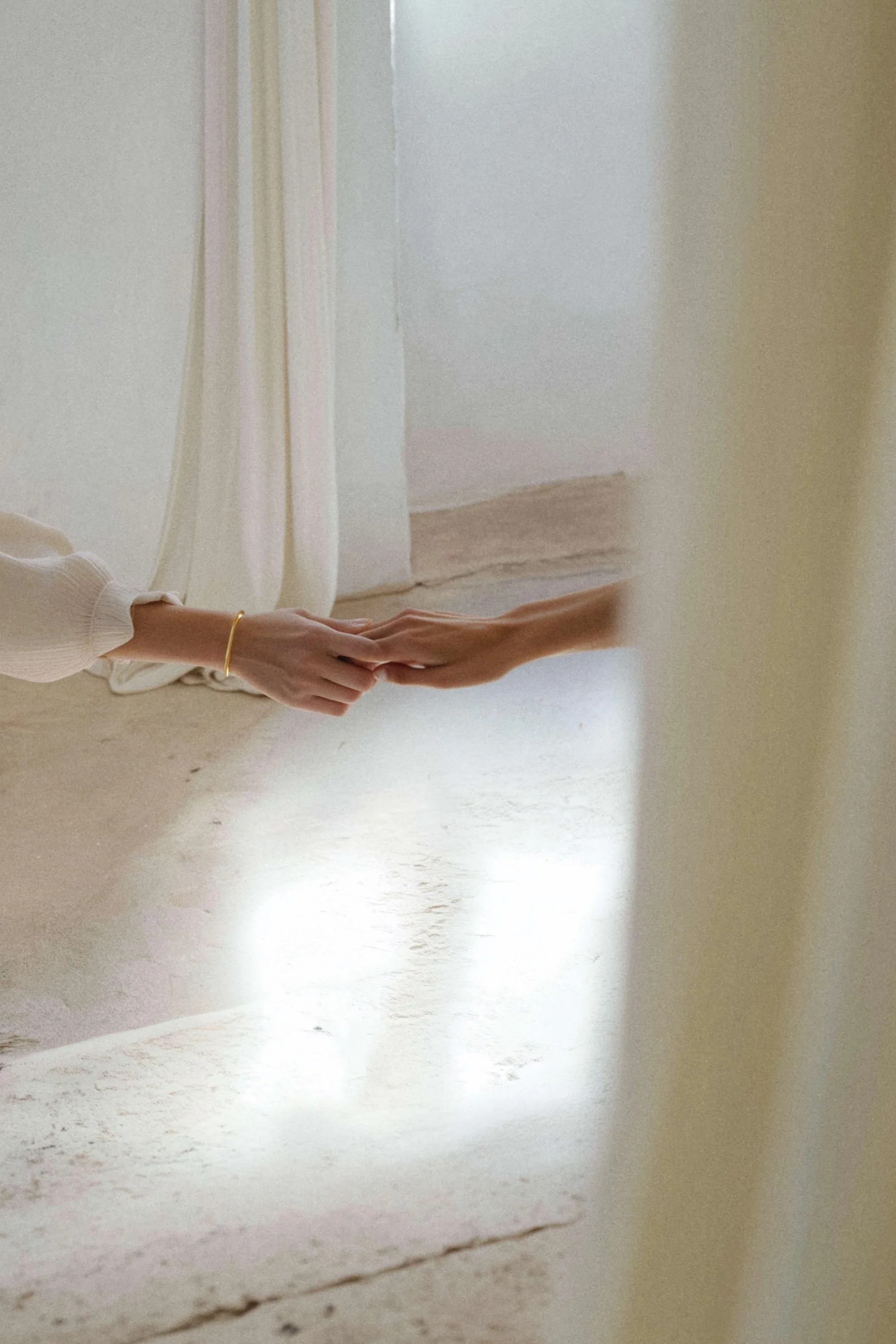 Two hands touching, one with a gold bracelet, in a softly lit, neutral-toned room.