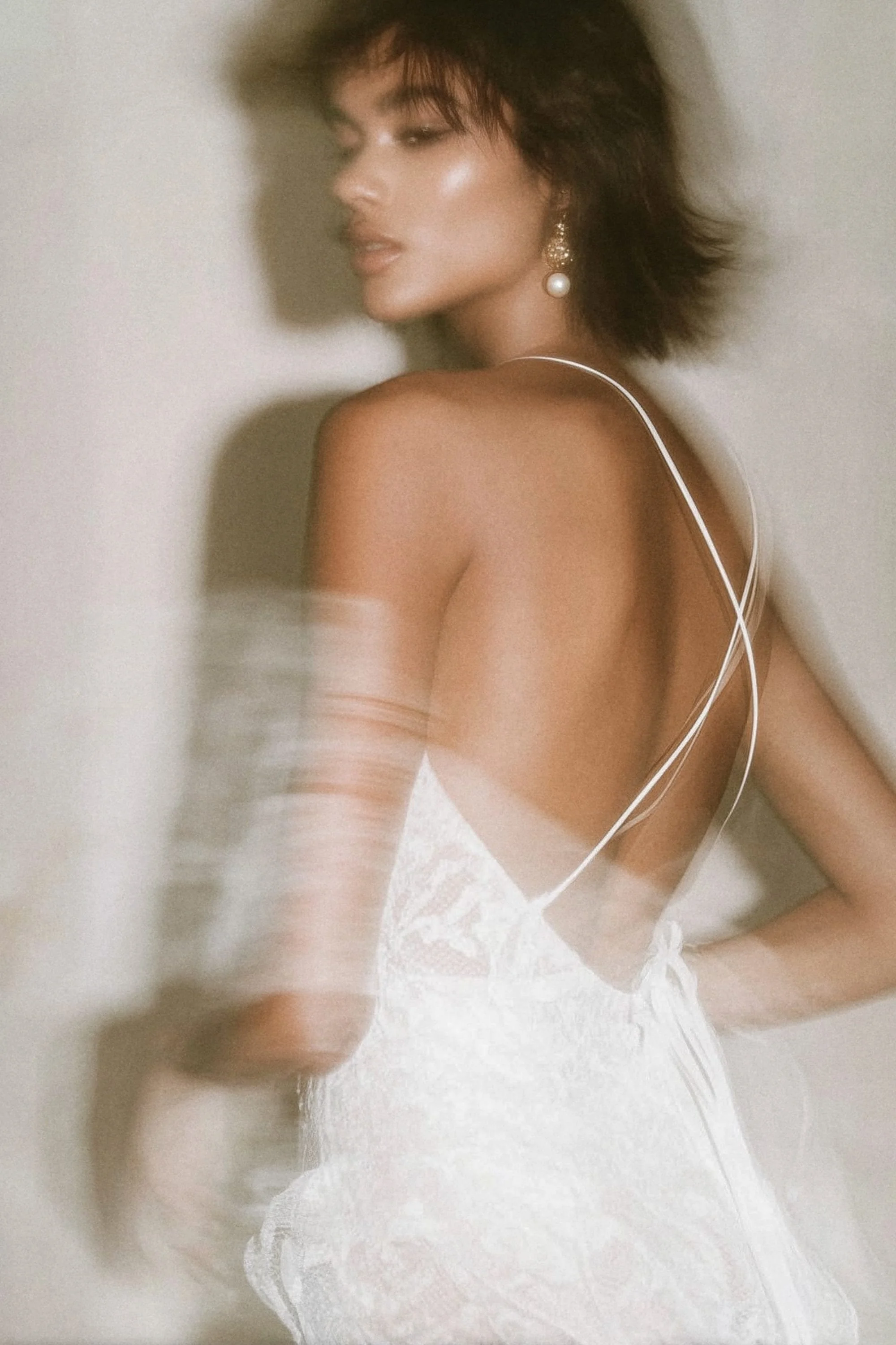 A woman in a white lace dress with thin straps, earrings, and short messy hair, standing against a plain background with a soft focus.