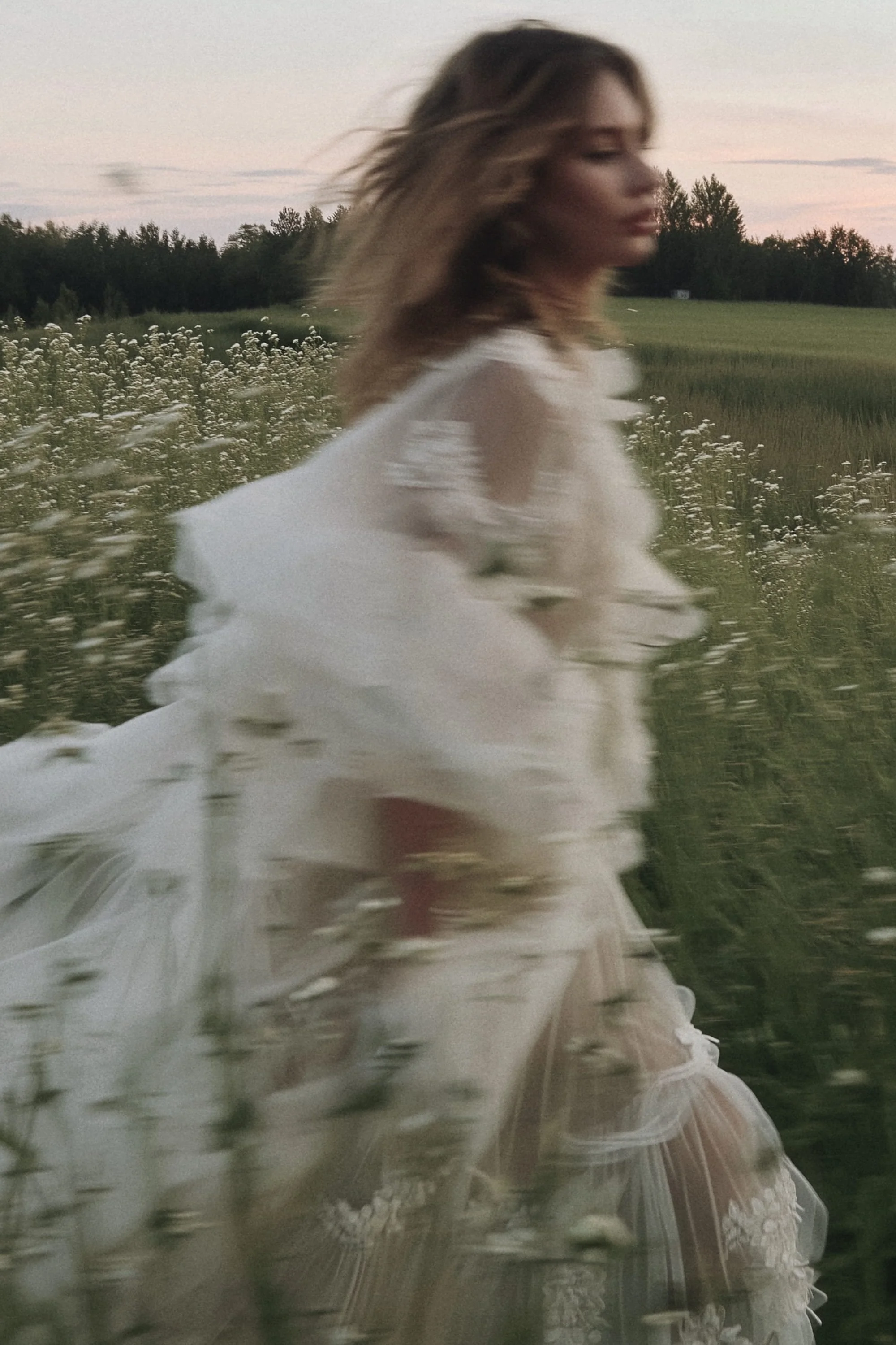A woman in a white dress walking through a field of wildflowers at dusk.