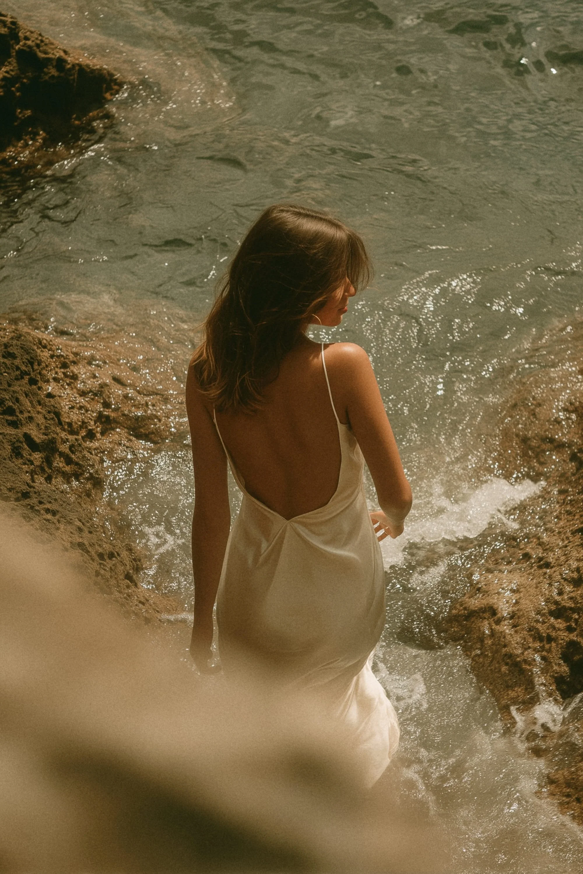 A woman in a beige dress standing in the water near rocks at a beach.
