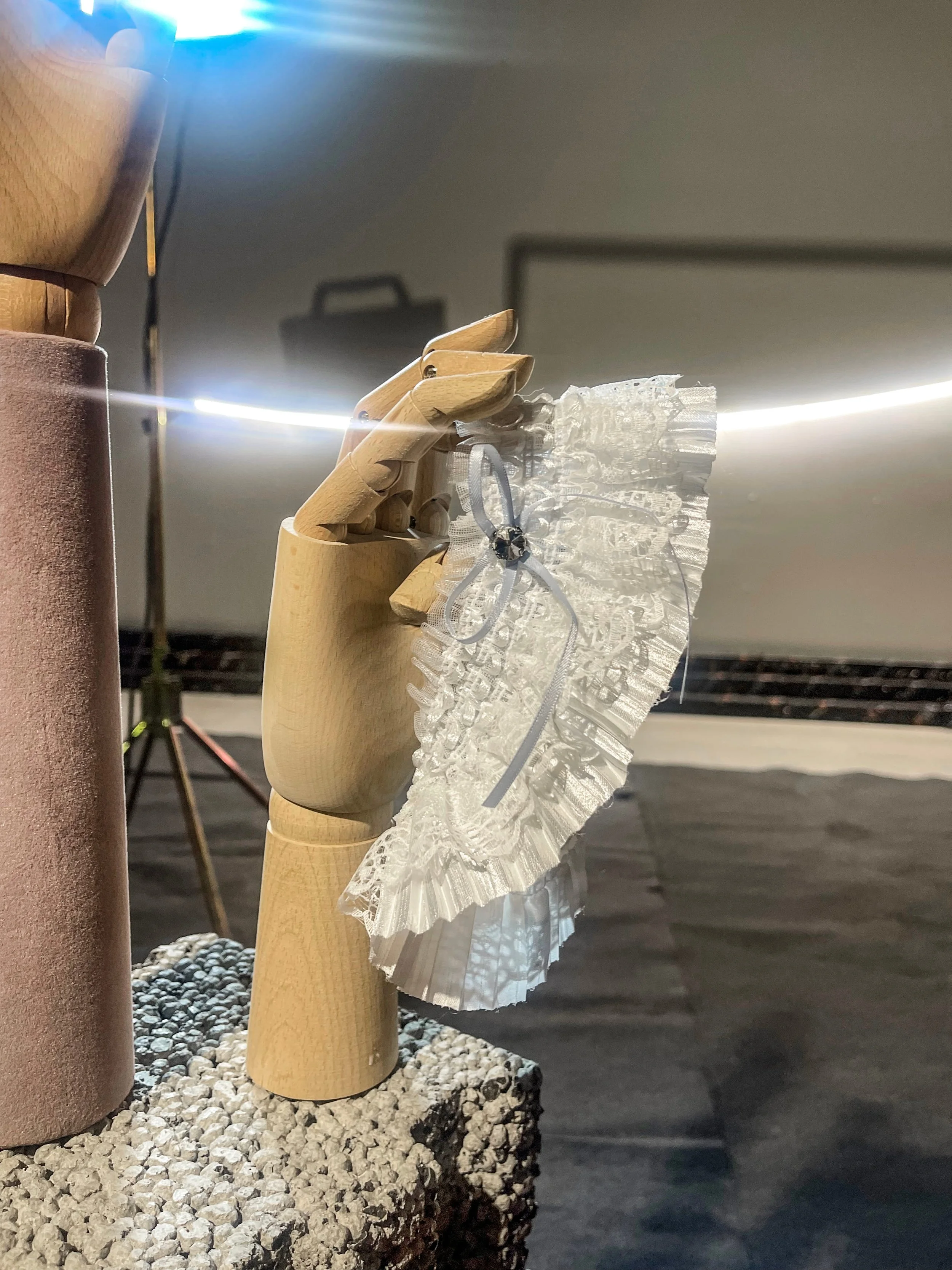 A wooden artist's hand model holding a white lace and ribbon bonnet decorated with a small bow and a rhinestone embellishment.