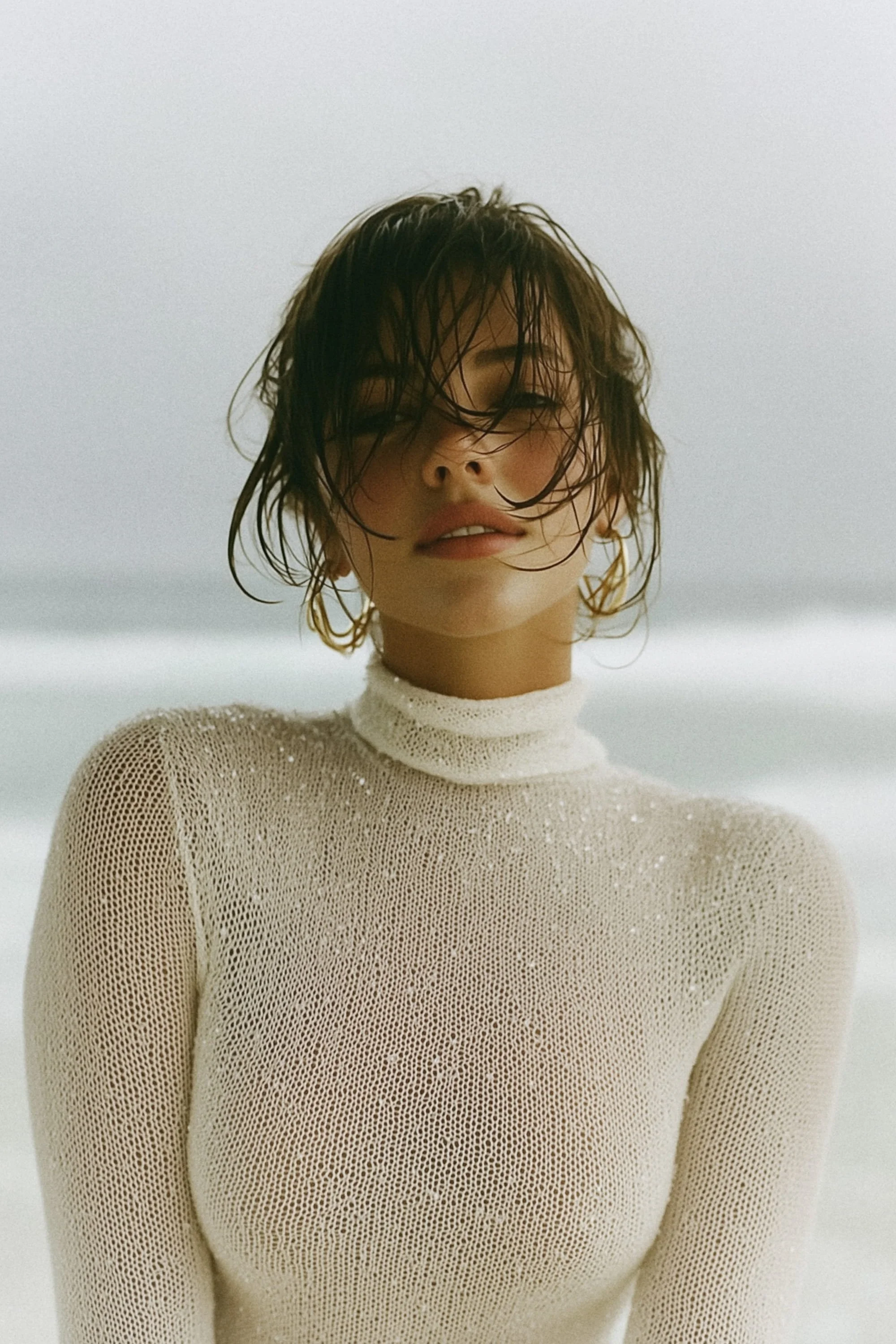 A woman with wet hair standing on a beach, wearing a cream-colored, long-sleeve, knitted turtleneck sweater.