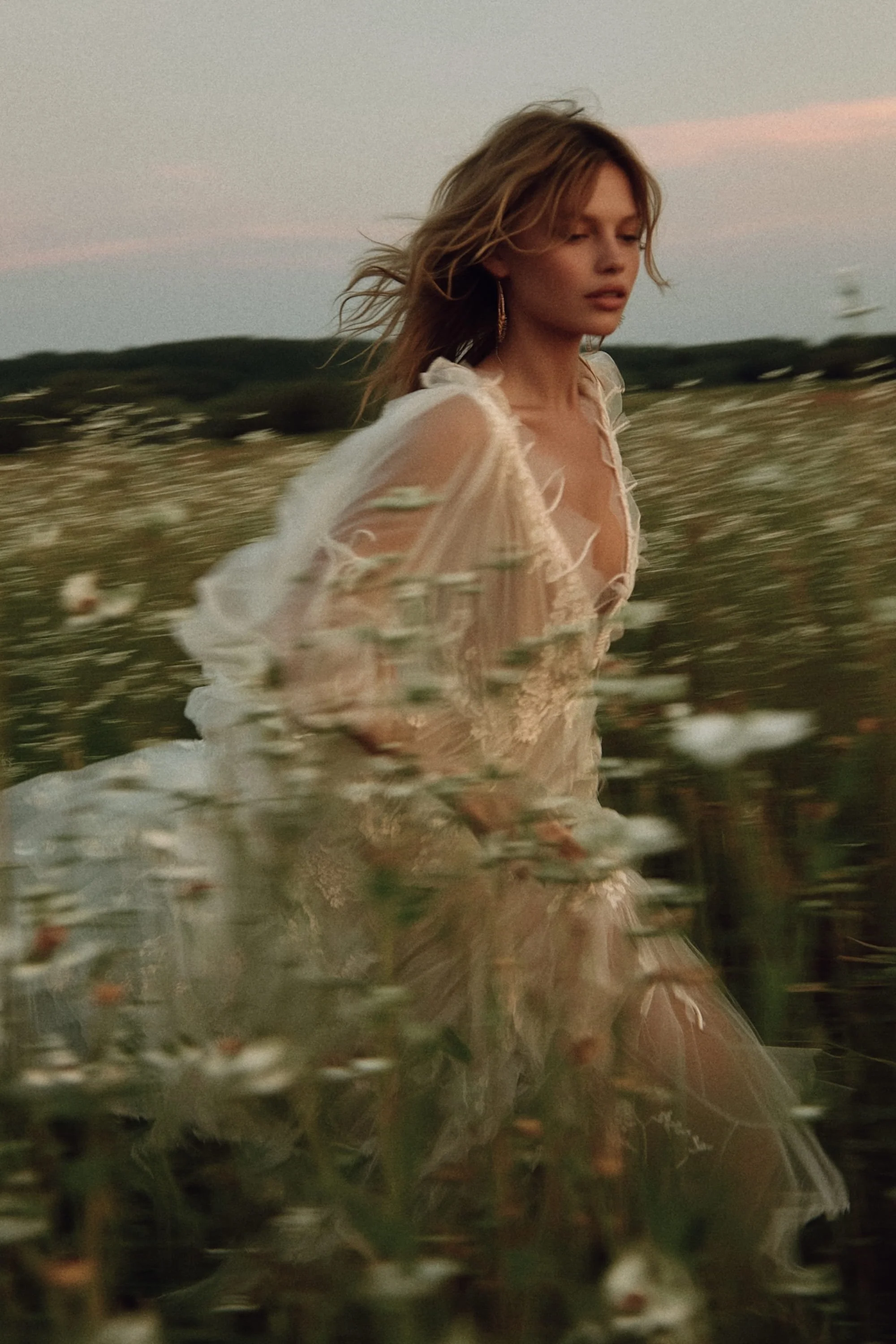 A woman in a sheer, lace dress walking through a field of flowers at sunset.