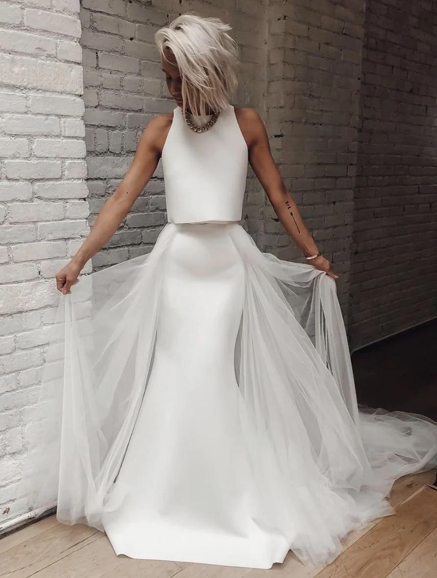 Woman wearing a white wedding gown, standing against a white brick wall, with her head tilted down and holding the sides of her dress.