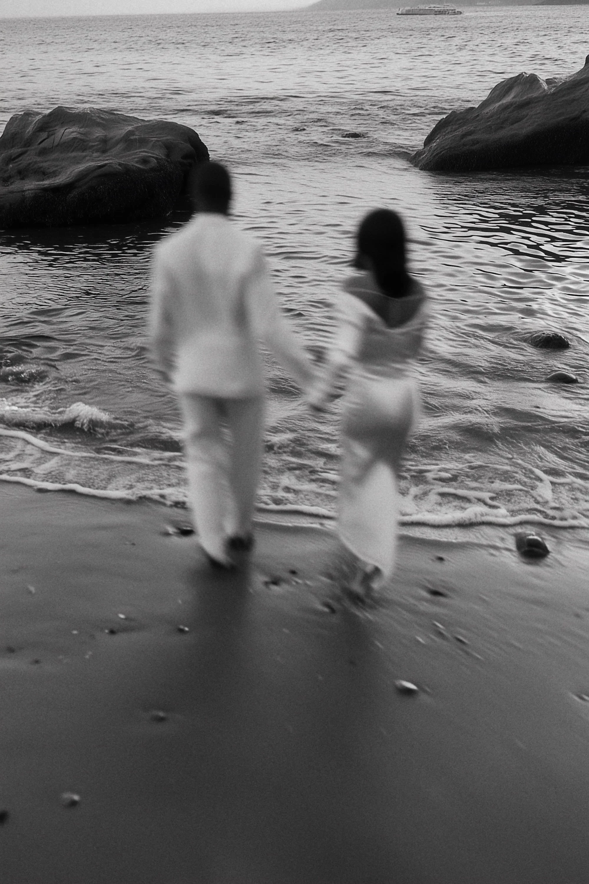 A black-and-white photo of a couple walking hand in hand along the shoreline, with large rocks in the water and the ocean in the background.