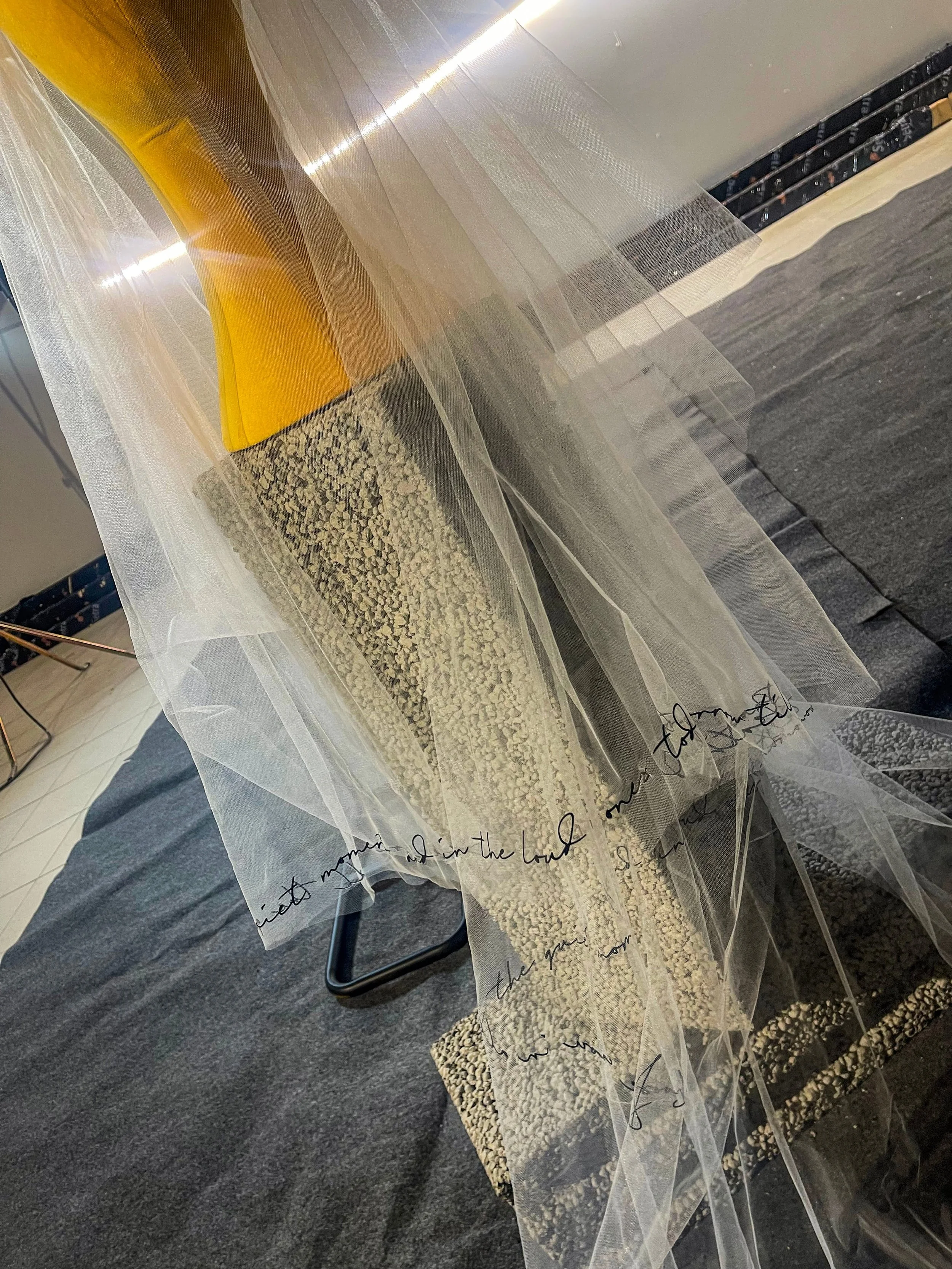 Decorative display featuring a yellow object and a textured base, draped with clear sheer fabric with handwritten black cursive writing.