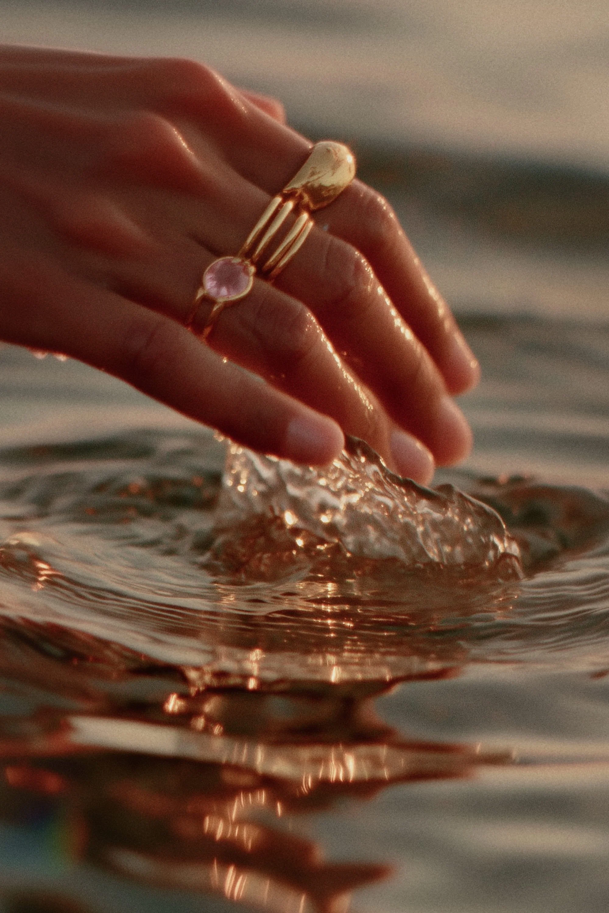 A hand with rings touching water in a lake or sea, creating ripples, with warm lighting.