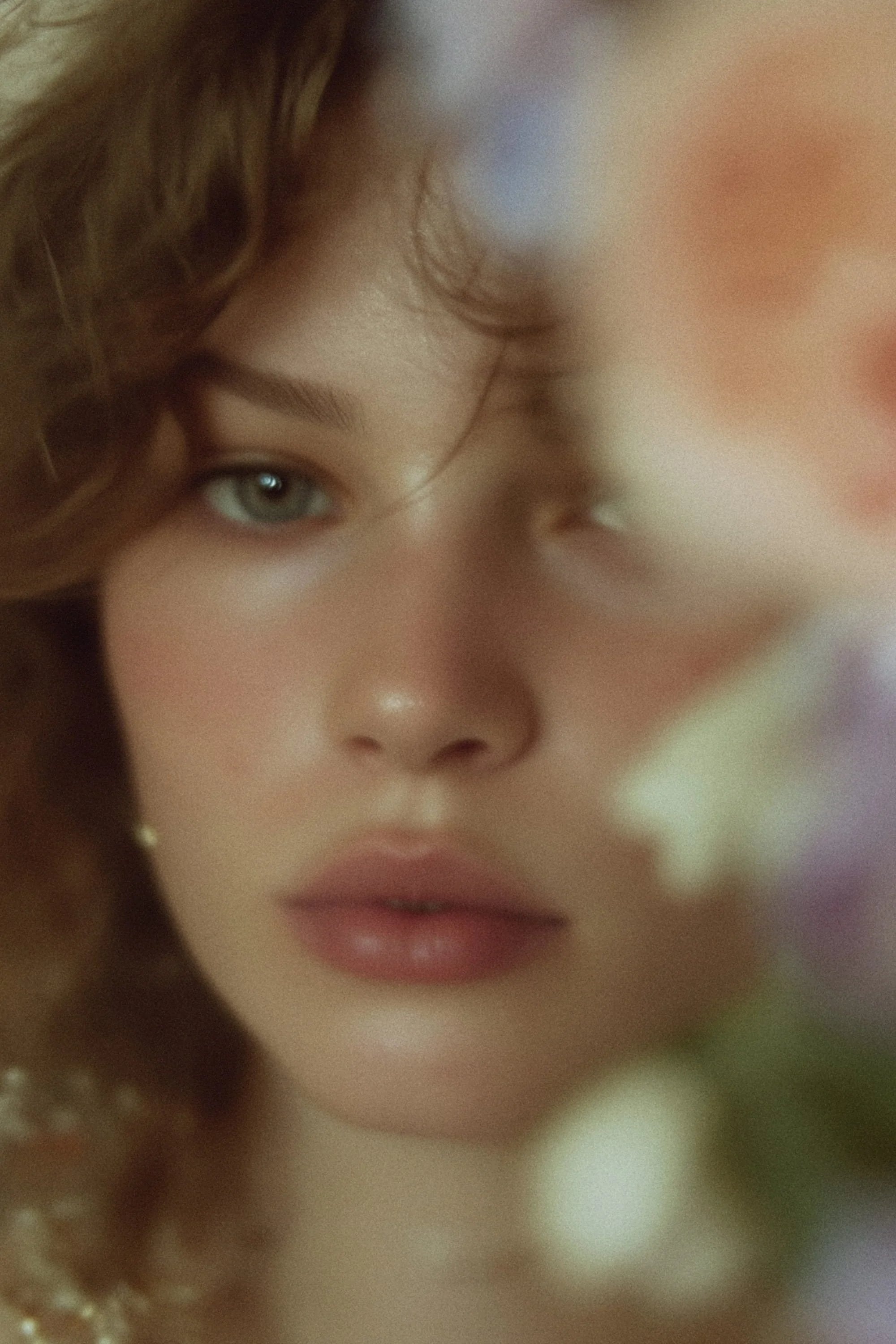 Close-up of a woman's face with light skin, curly hair, and blue eyes, partially obscured by colorful out-of-focus flowers.