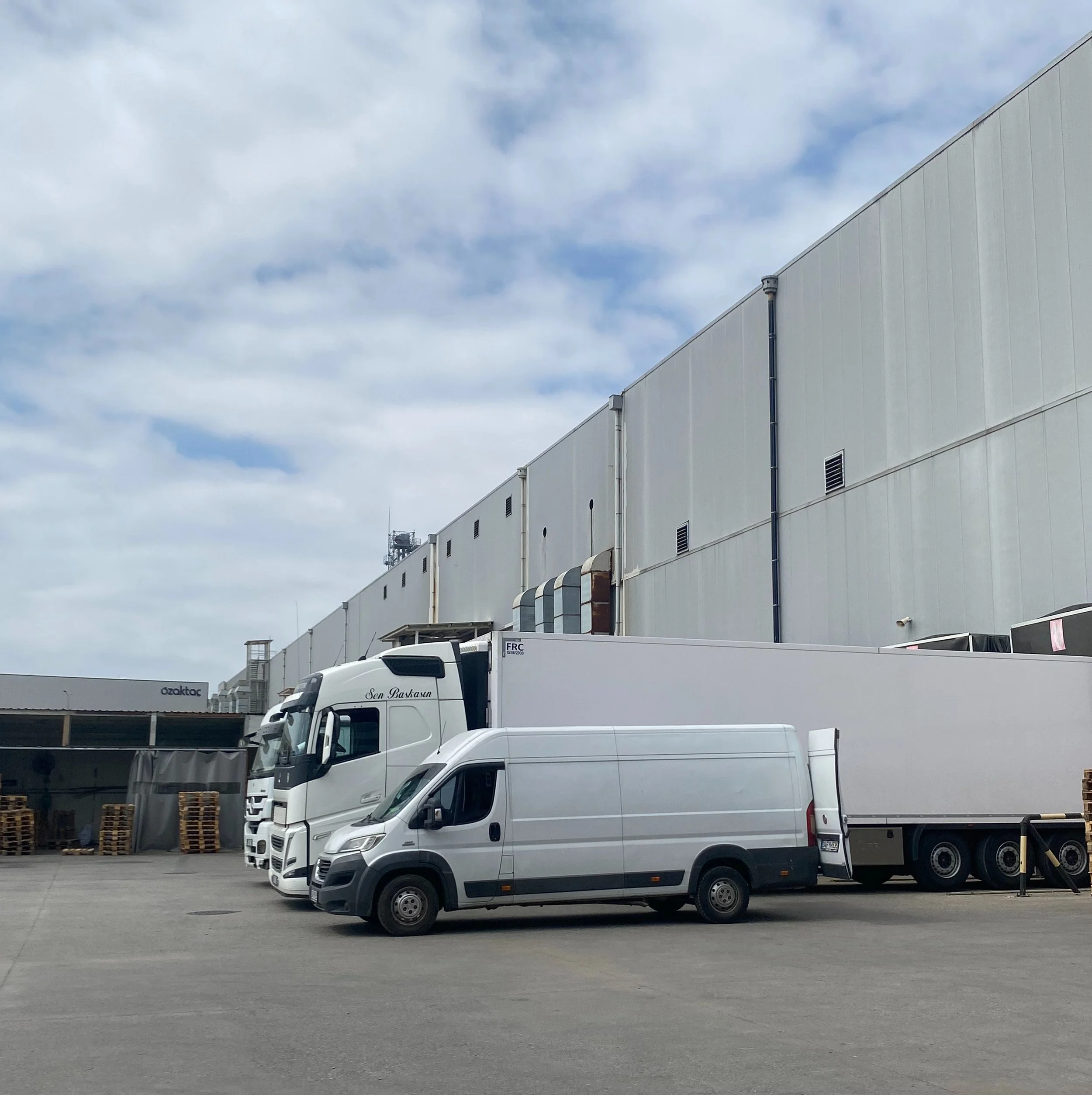 Parking lot with two white trucks, a smaller white van, and a large warehouse building under a partly cloudy sky.