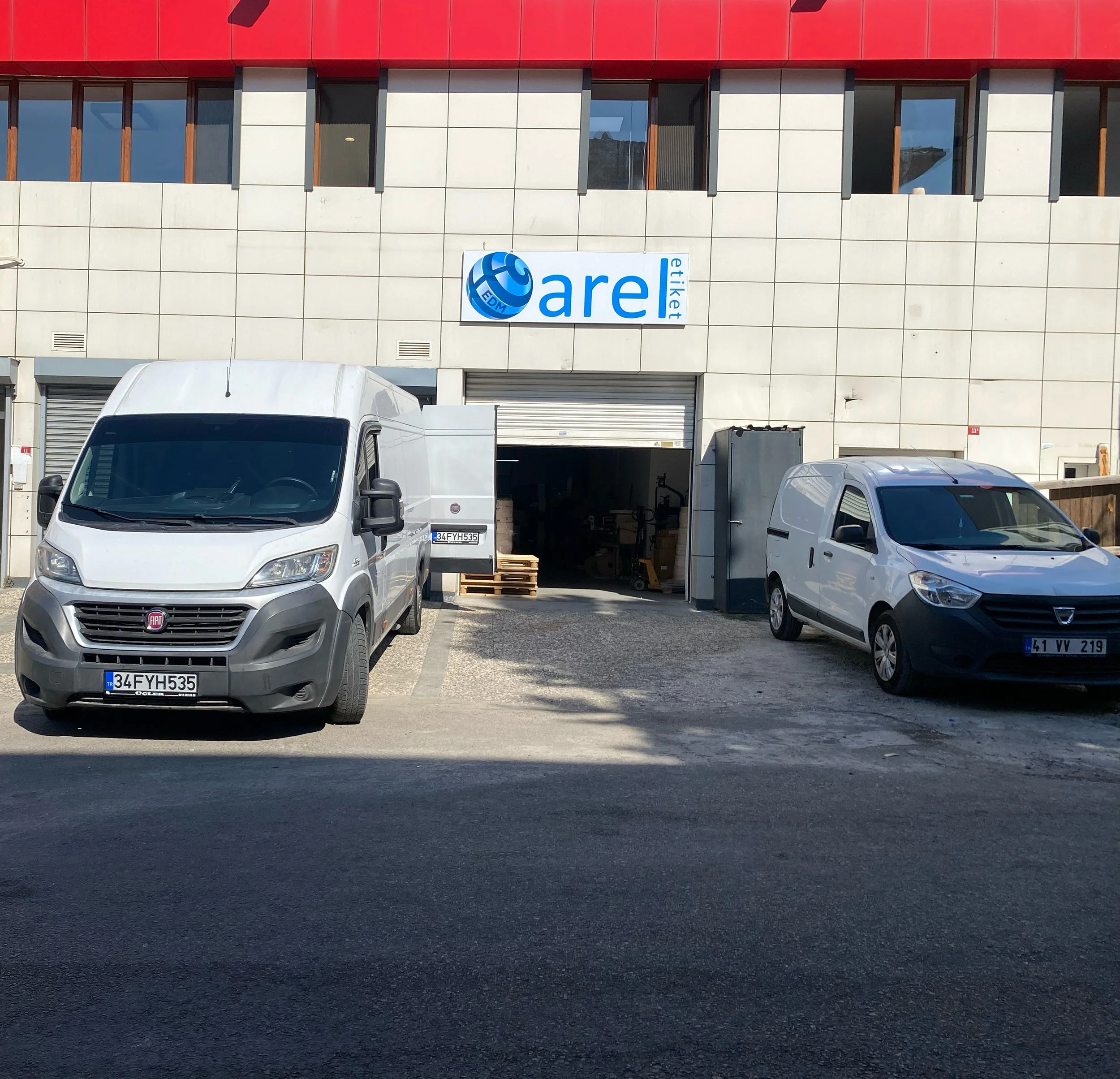 Front view of a warehouse with two parked white vans in front, open loading dock with some pallets and industrial equipment inside, and a sign reading 'are etiket' above the entrance.