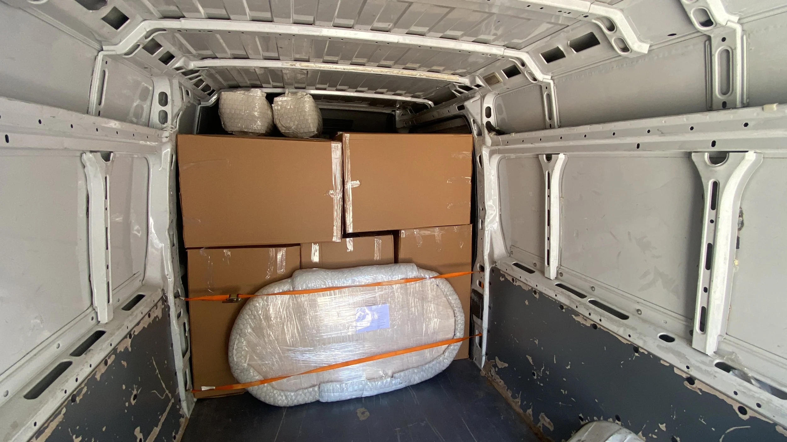 Interior of a cargo van loaded with cardboard boxes and a wrapped item secured with orange straps, with additional packages in the back.