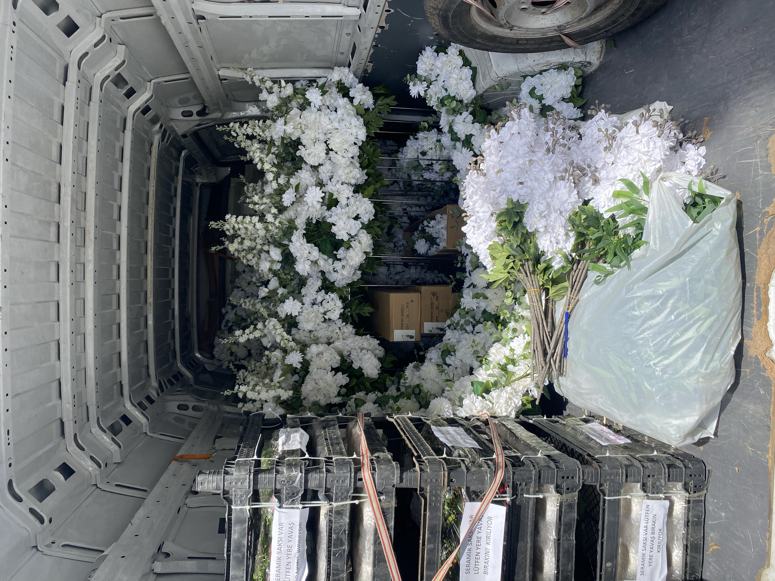 A collection of white flowers, including hydrangeas and other blooms, arranged and placed inside the back of a van. Some flowers are bundled with rubber bands, and there are additional flower arrangements and boxes nearby.