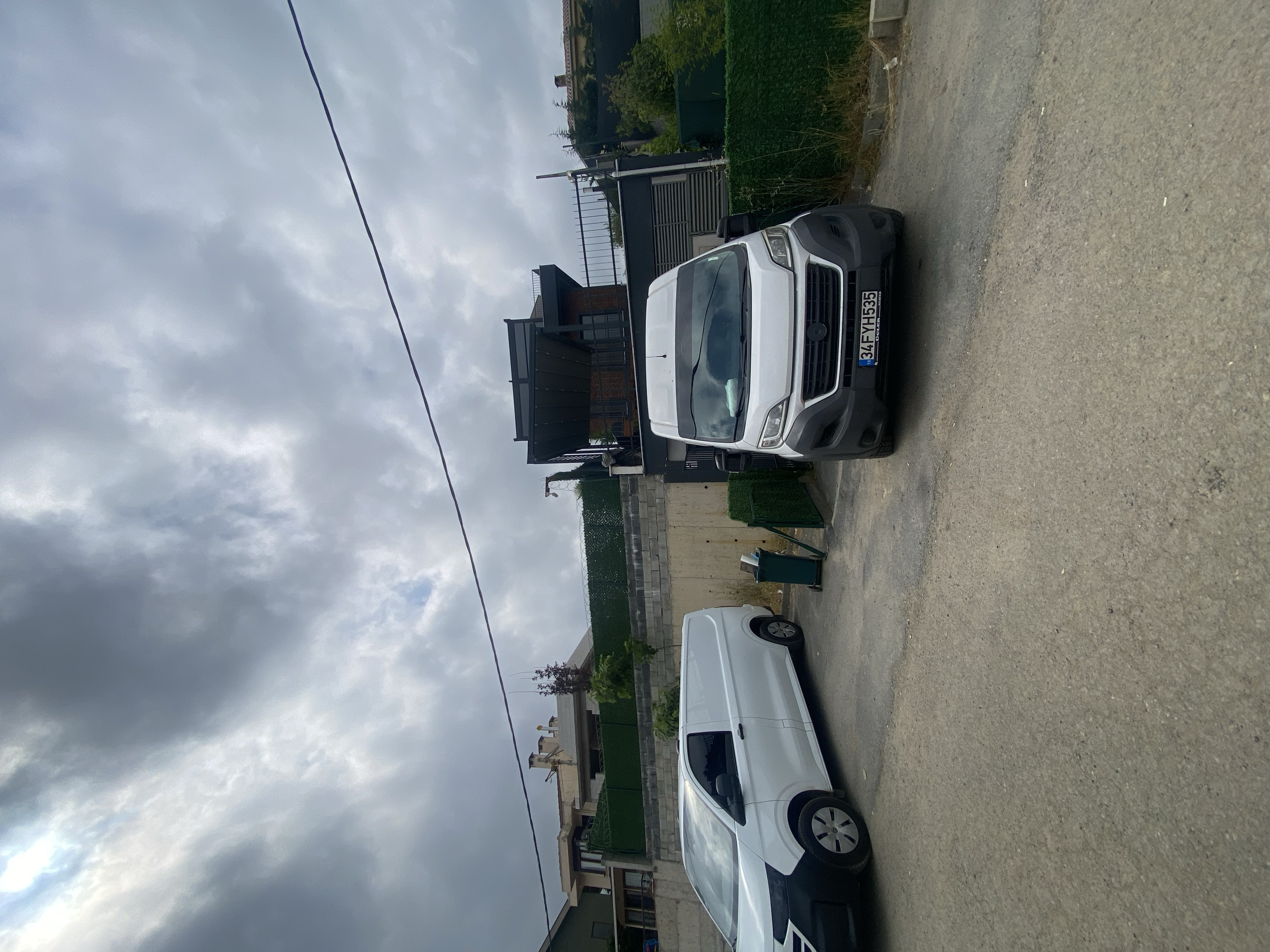 A metallic gray pickup truck with a black front bumper and grille parked in front of a house with a balcony, on a gravel driveway. Two white vehicles are also visible, one parked behind the pickup and another to the side. Overcast sky with gray cloud