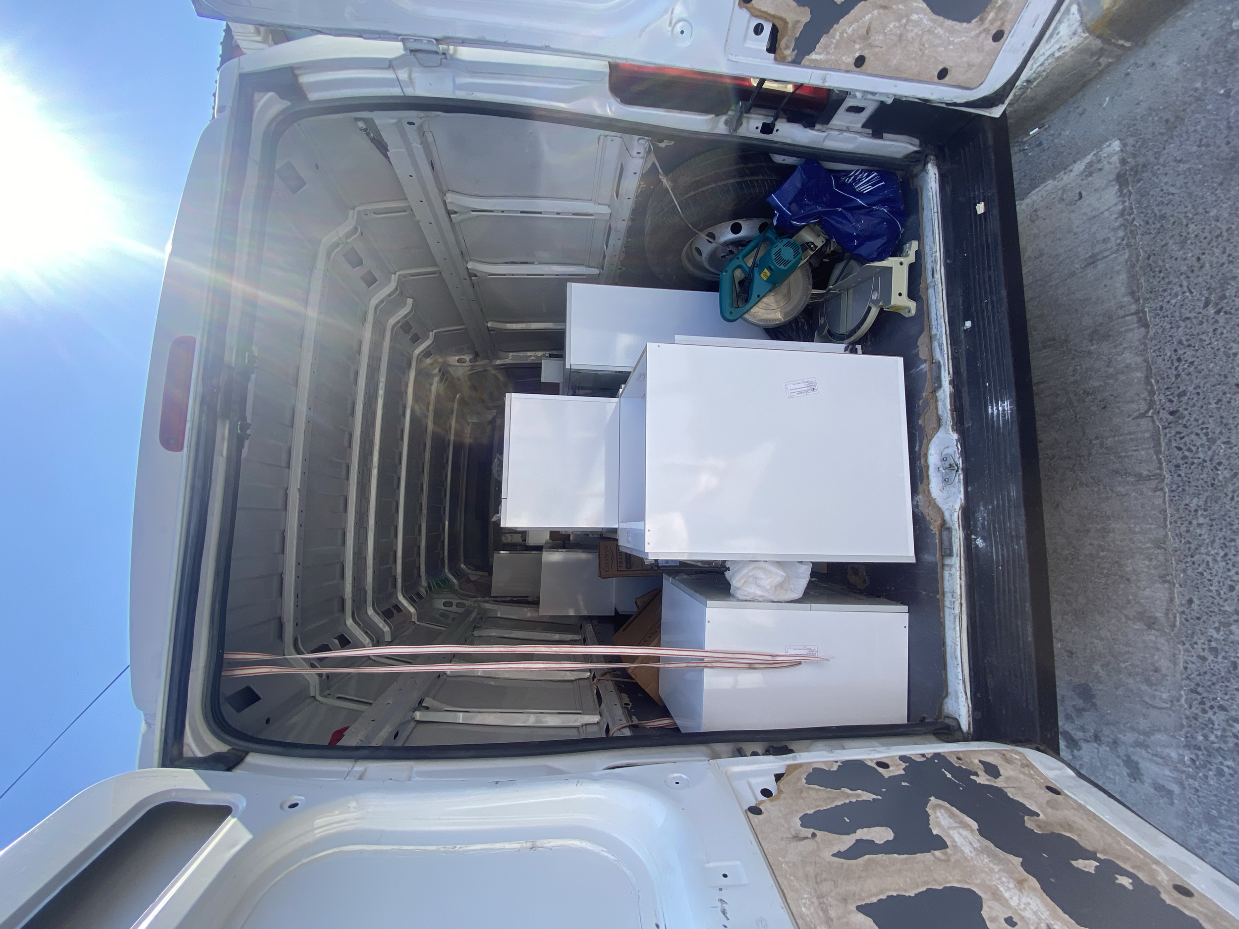 The back of a white delivery van with its doors open, revealing various household furniture and items including a small white refrigerator, a small white cabinet, a cardboard box, and some tools. The van shows signs of wear and peeling paint on the i