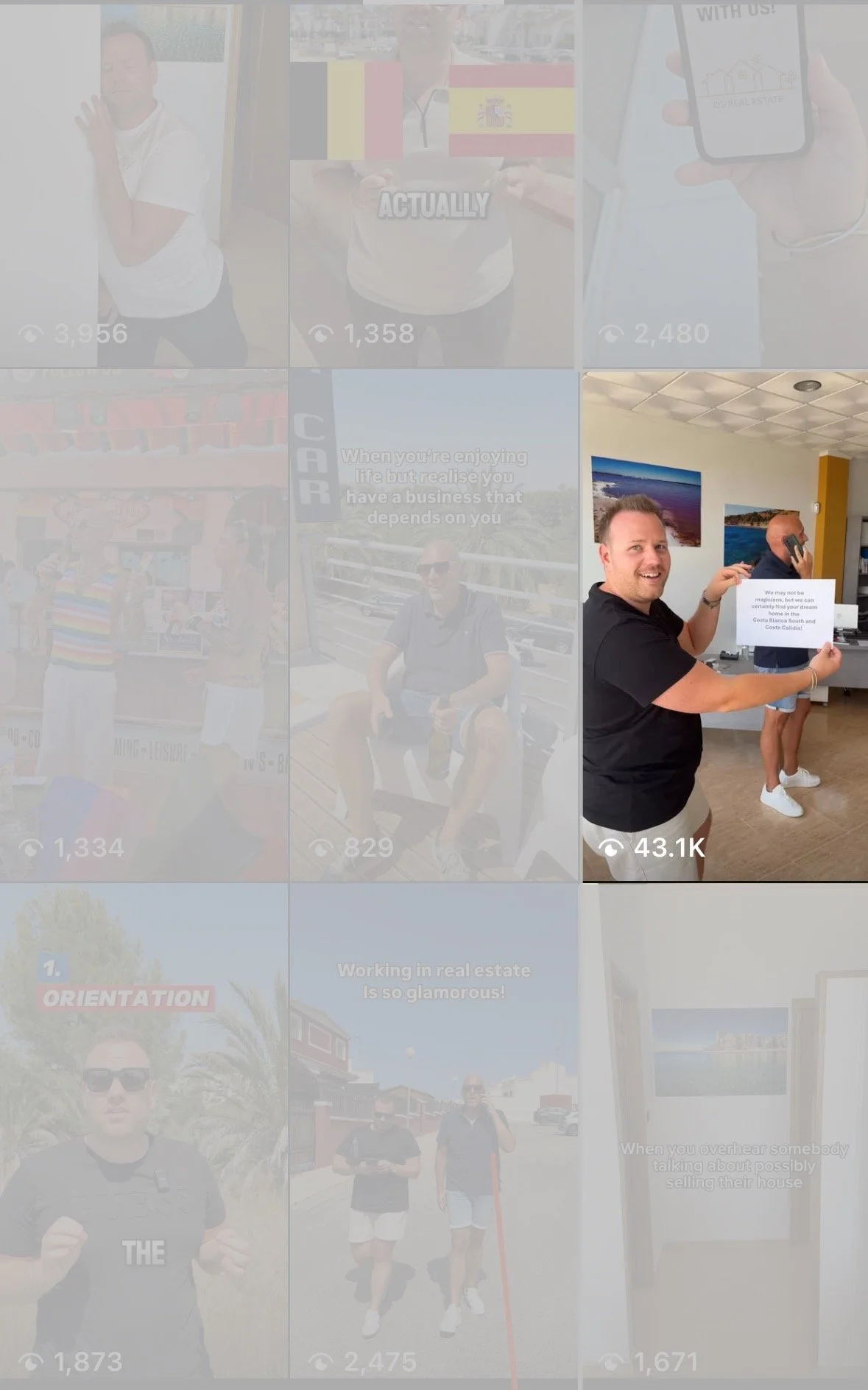 A man smiling and holding a sign in an office, with a woman talking on the phone in the background.