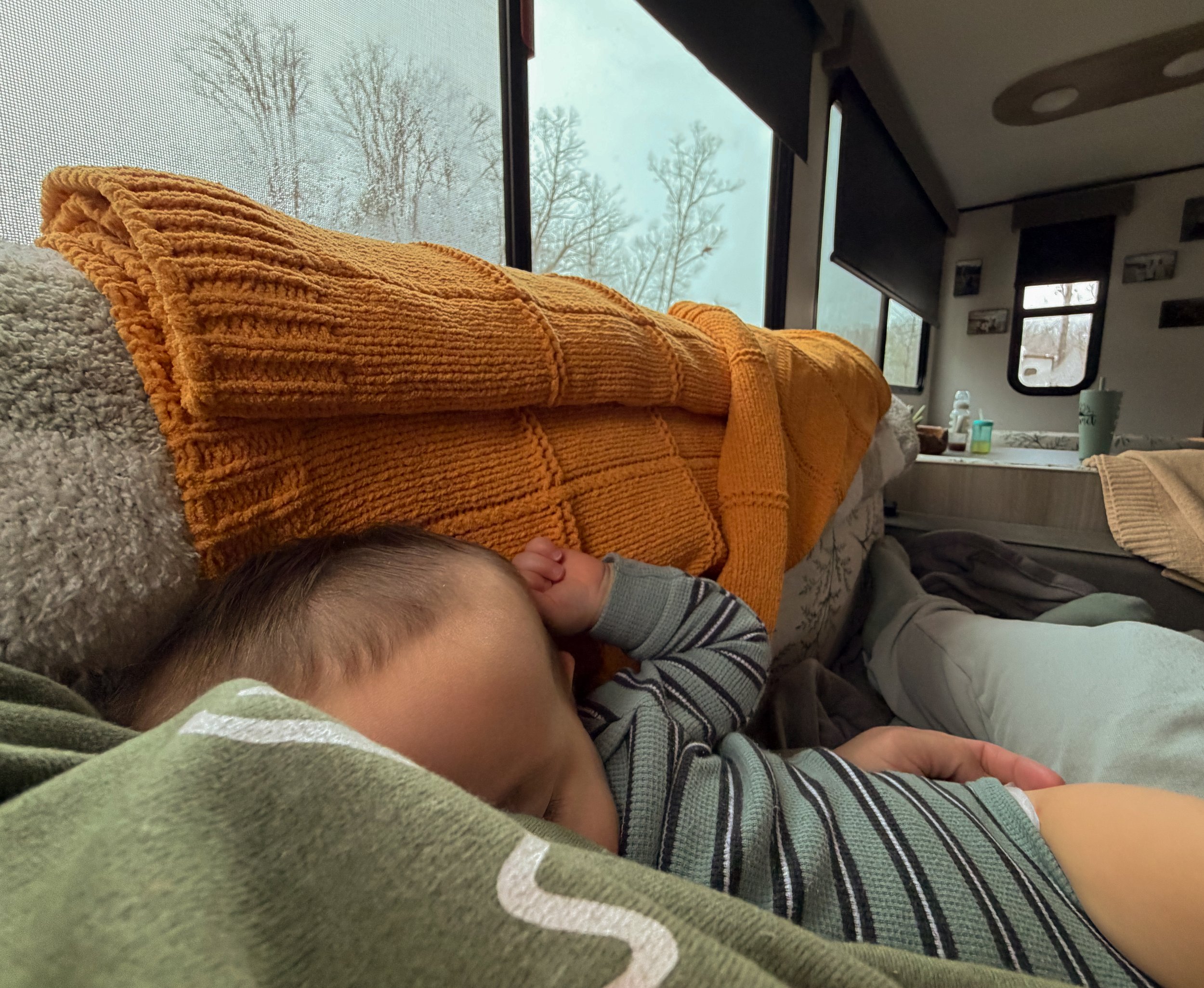 Sleeping baby inside a warm RV camper during a winter storm, with sleet-covered trees visible through the window, showing cozy off-grid family life.
