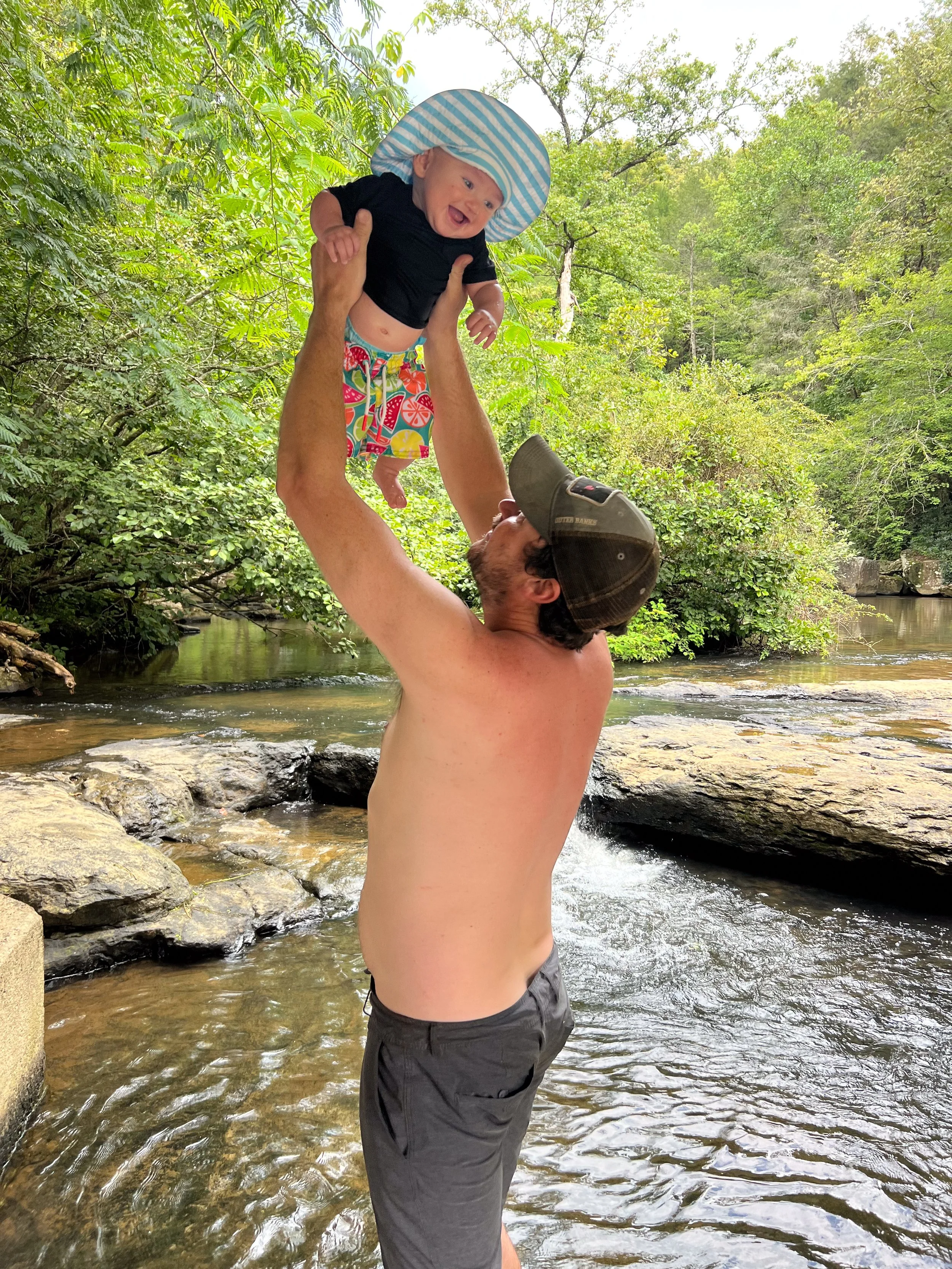 Father lifting his baby in a creek surrounded by nature, representing family, adventure, and raising a child outdoors.