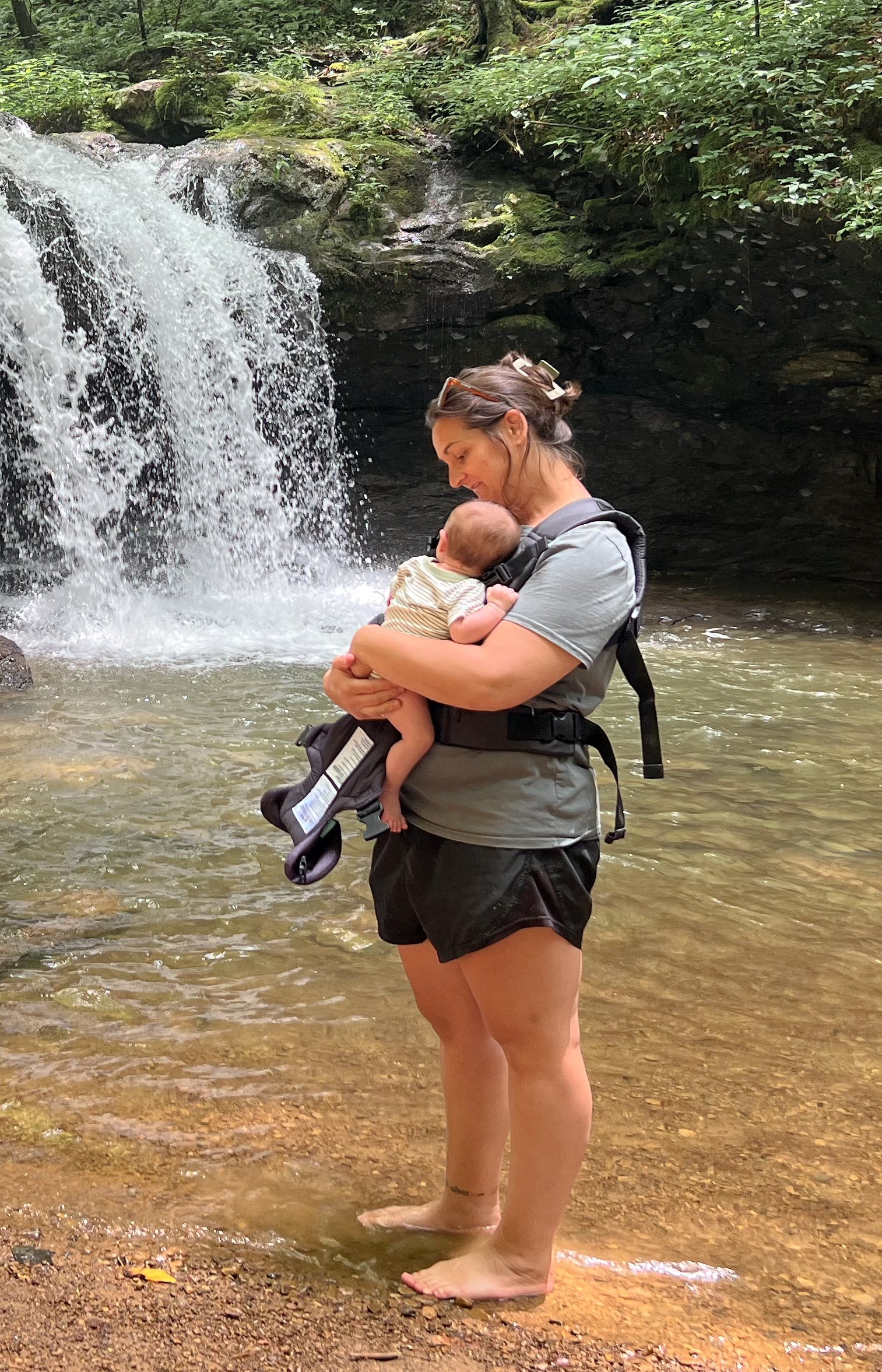 A mother standing barefoot in a shallow creek, holding her infant close while looking down at him, with a soft waterfall in the background — a quiet moment of presence and everyday life in nature.