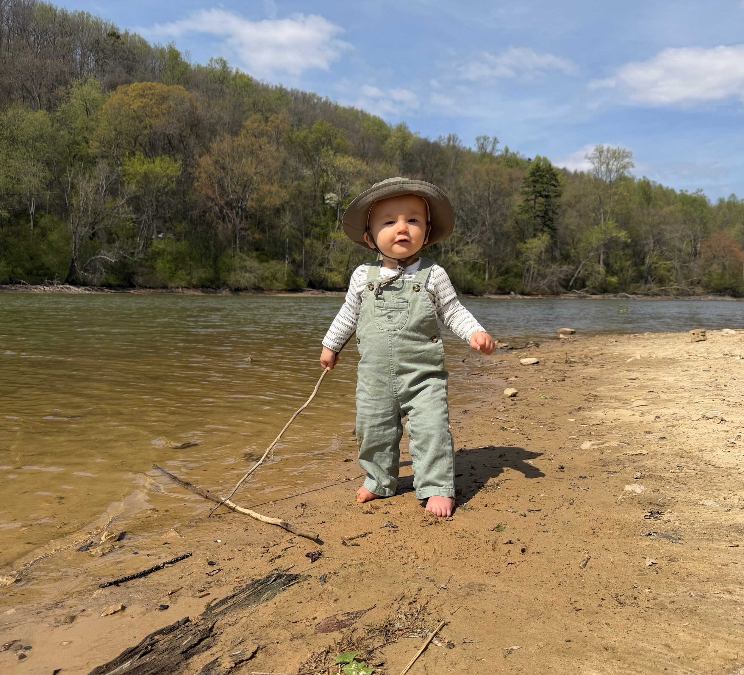 One-year-old exploring a sandy riverbank barefoot while holding a walking stick outdoors.