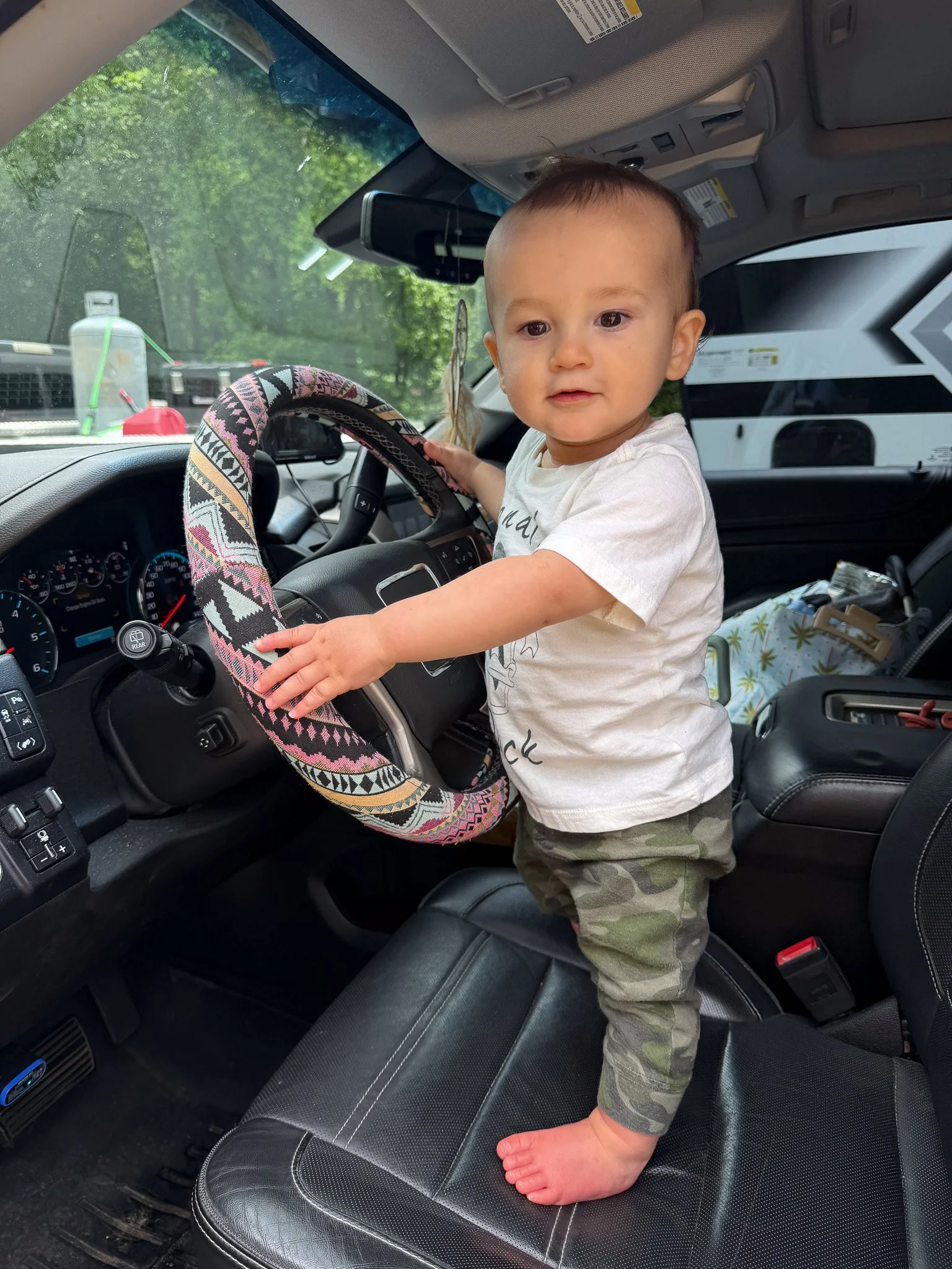 One-year-old Banks standing at the steering wheel of the family SUV with the travel trailer visible outside.