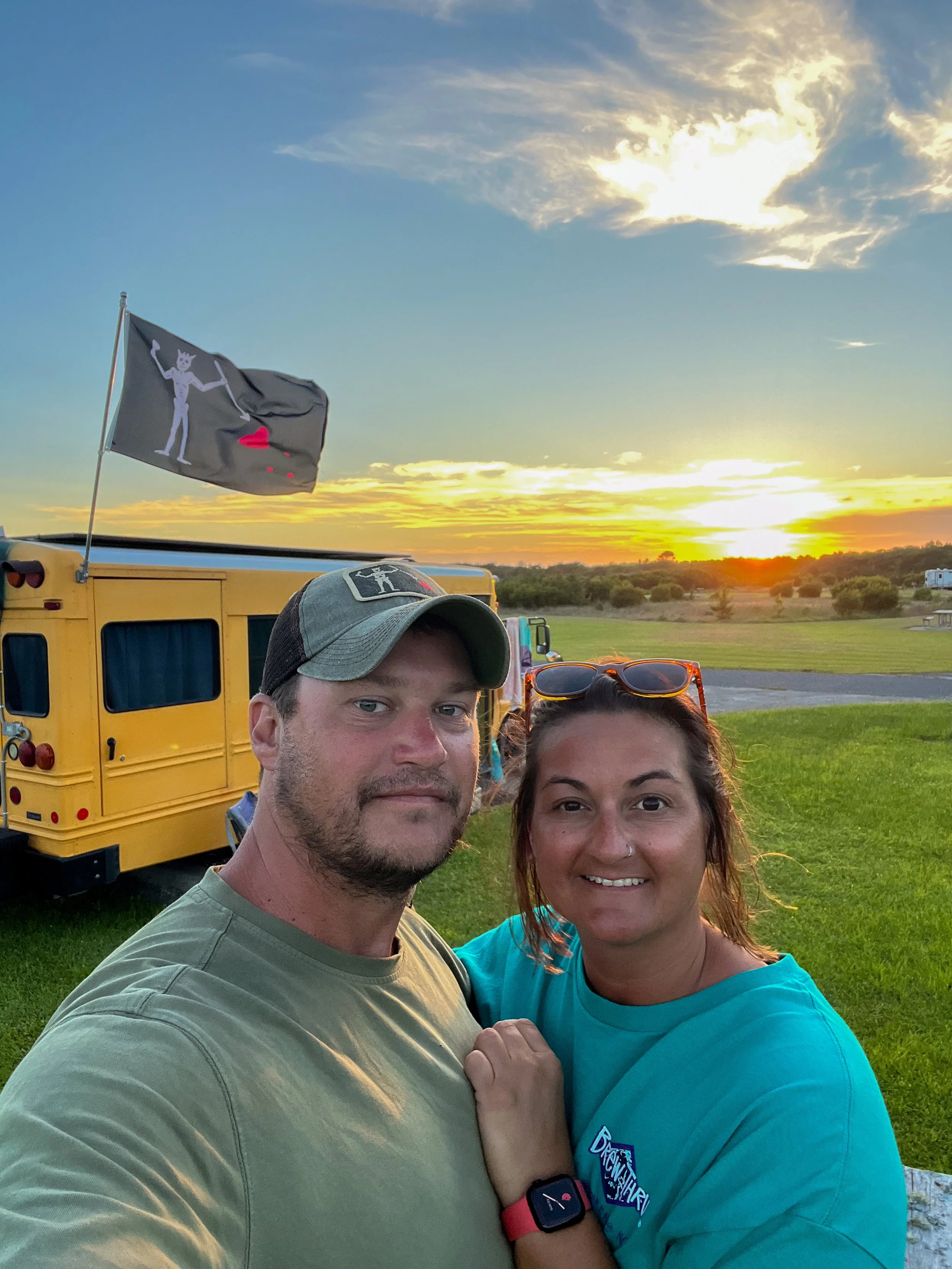 Couple standing in front of a converted school bus at sunset, representing long-term nomadic living and life on the road.