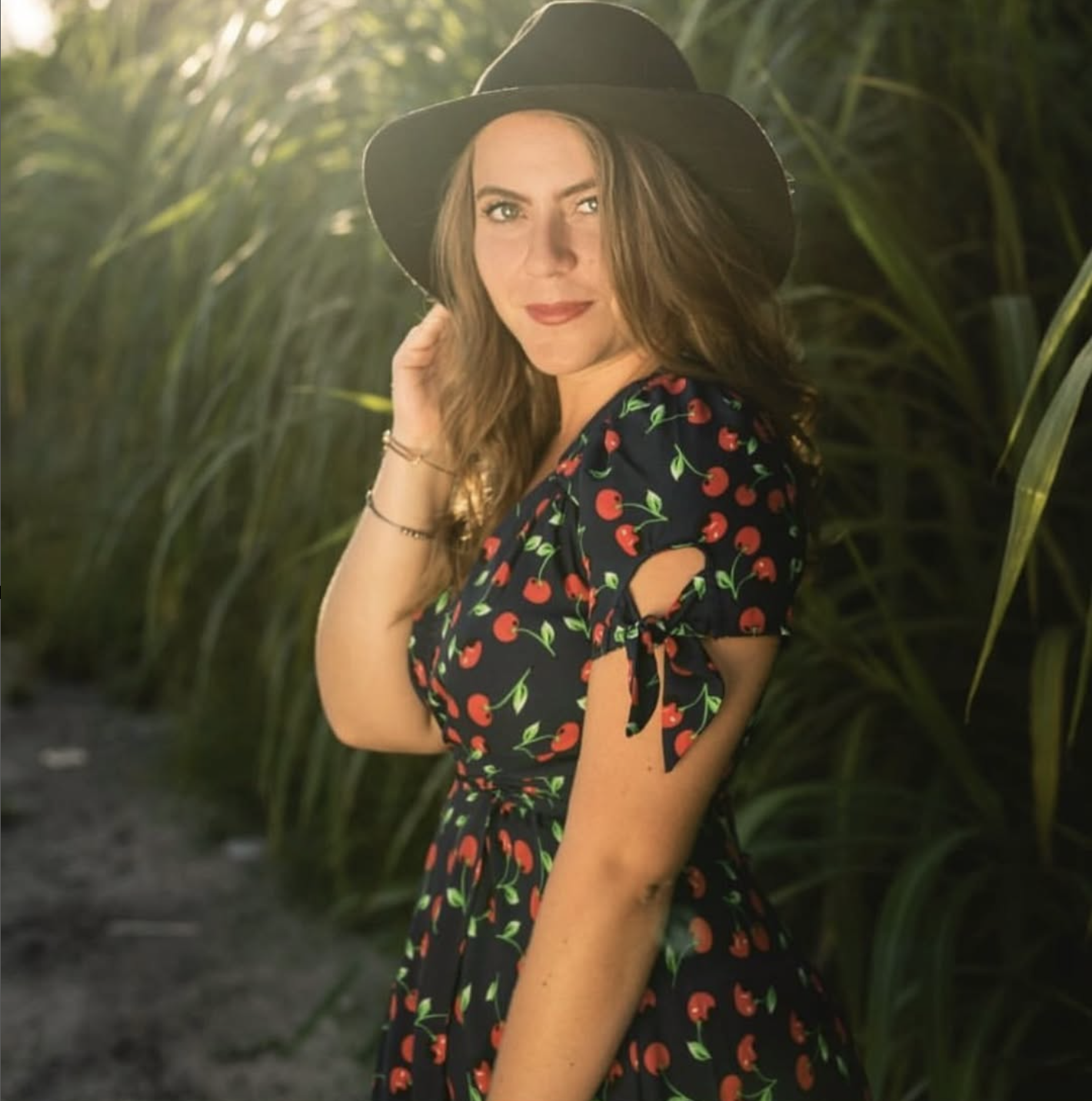 A woman wearing a black hat and a dark dress with a cherry pattern, standing outdoors in front of tall green foliage, looking at the camera.