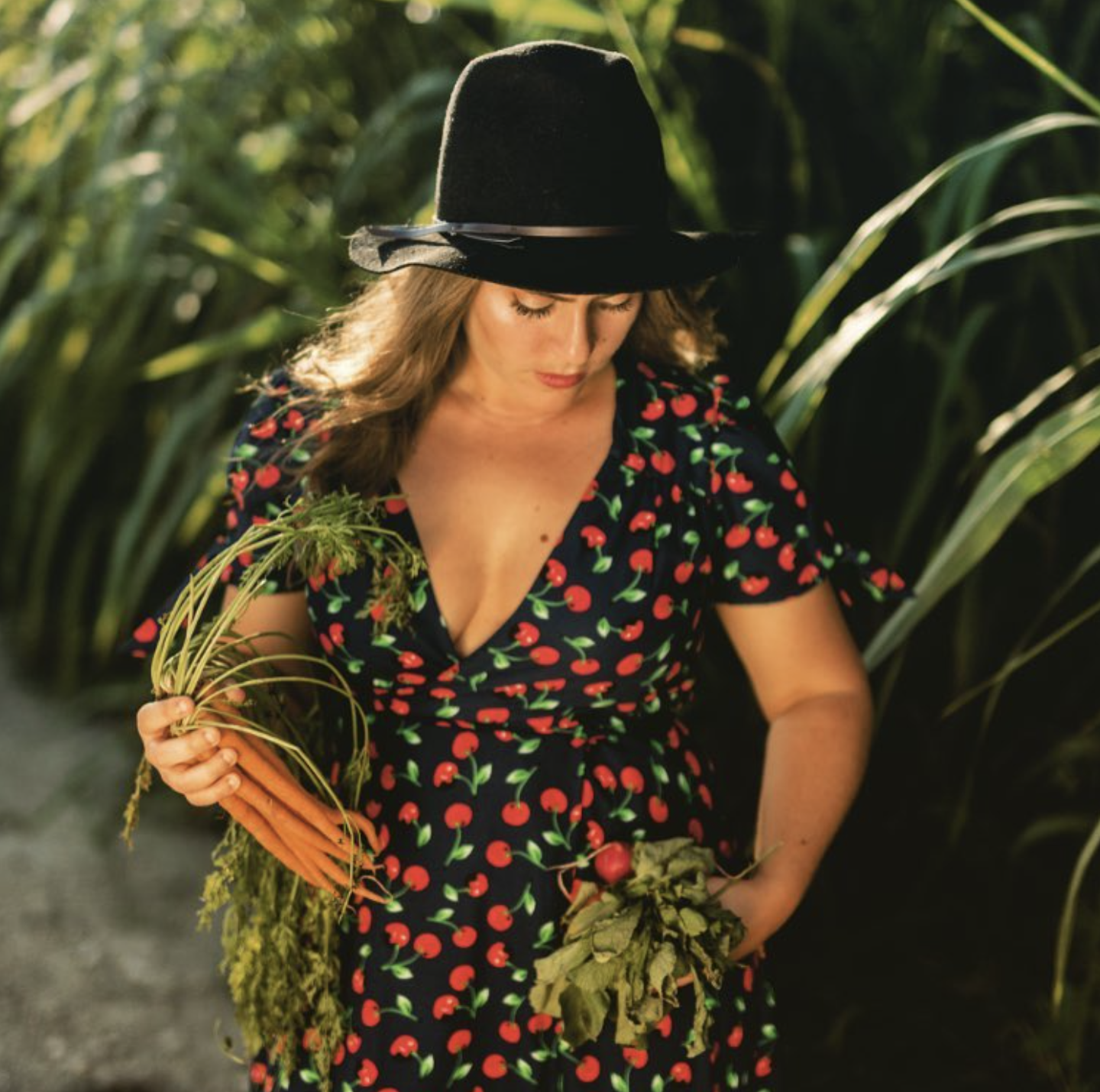 A woman wearing a black hat and a cherry-patterned dress holds a bunch of carrots and green leafy vegetables in a lush, sunny agricultural field.