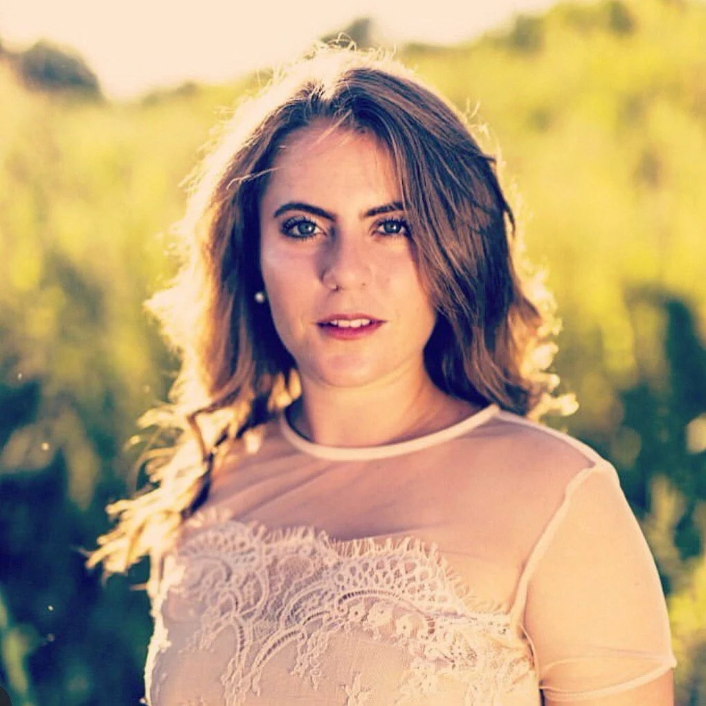 A woman with shoulder-length brown hair and blue eyes, wearing a sheer lace top, stands outdoors in a sunlit field with green foliage in the background.