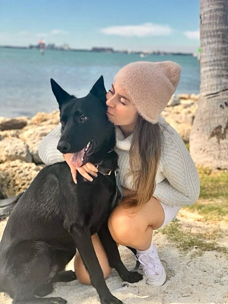 A woman with long brown hair wearing a beige beanie, white sweater, and white sneakers is hugging and kissing a black German Shepherd dog on a beach with rocks, sand, a palm tree, and the ocean in the background.