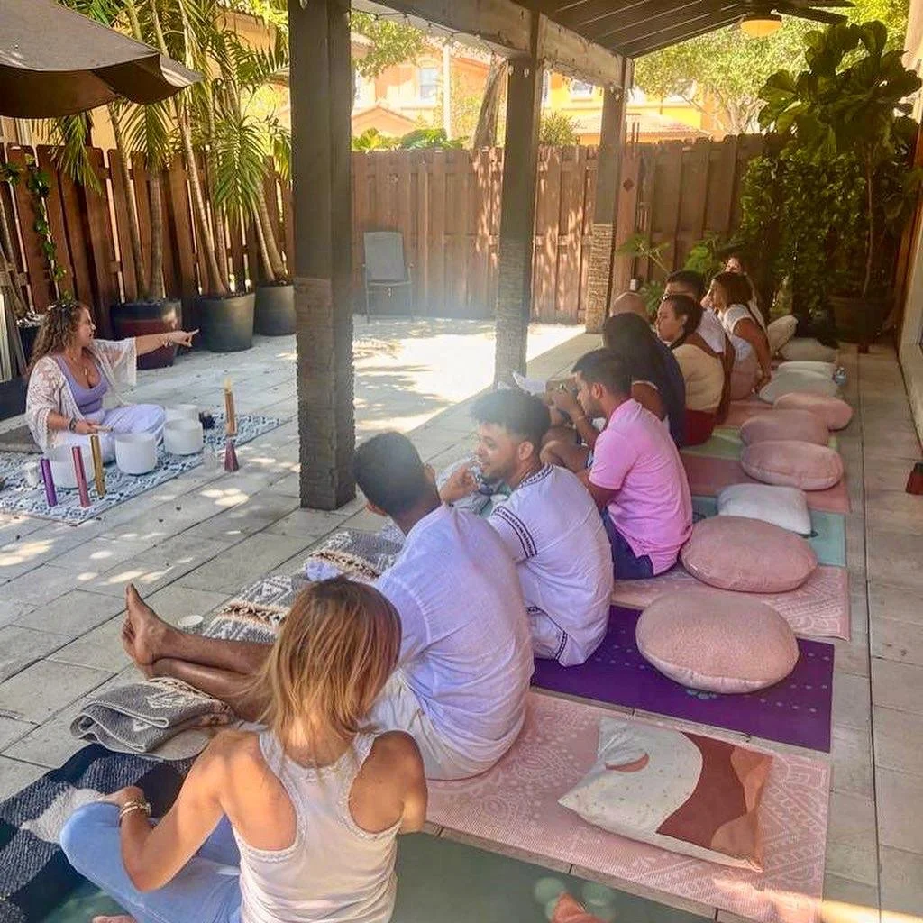 A woman leading a yoga or meditation class outdoors, with participants sitting on mats and cushions, sitting cross-legged and facing her in a shaded patio area surrounded by plants and wooden fencing.