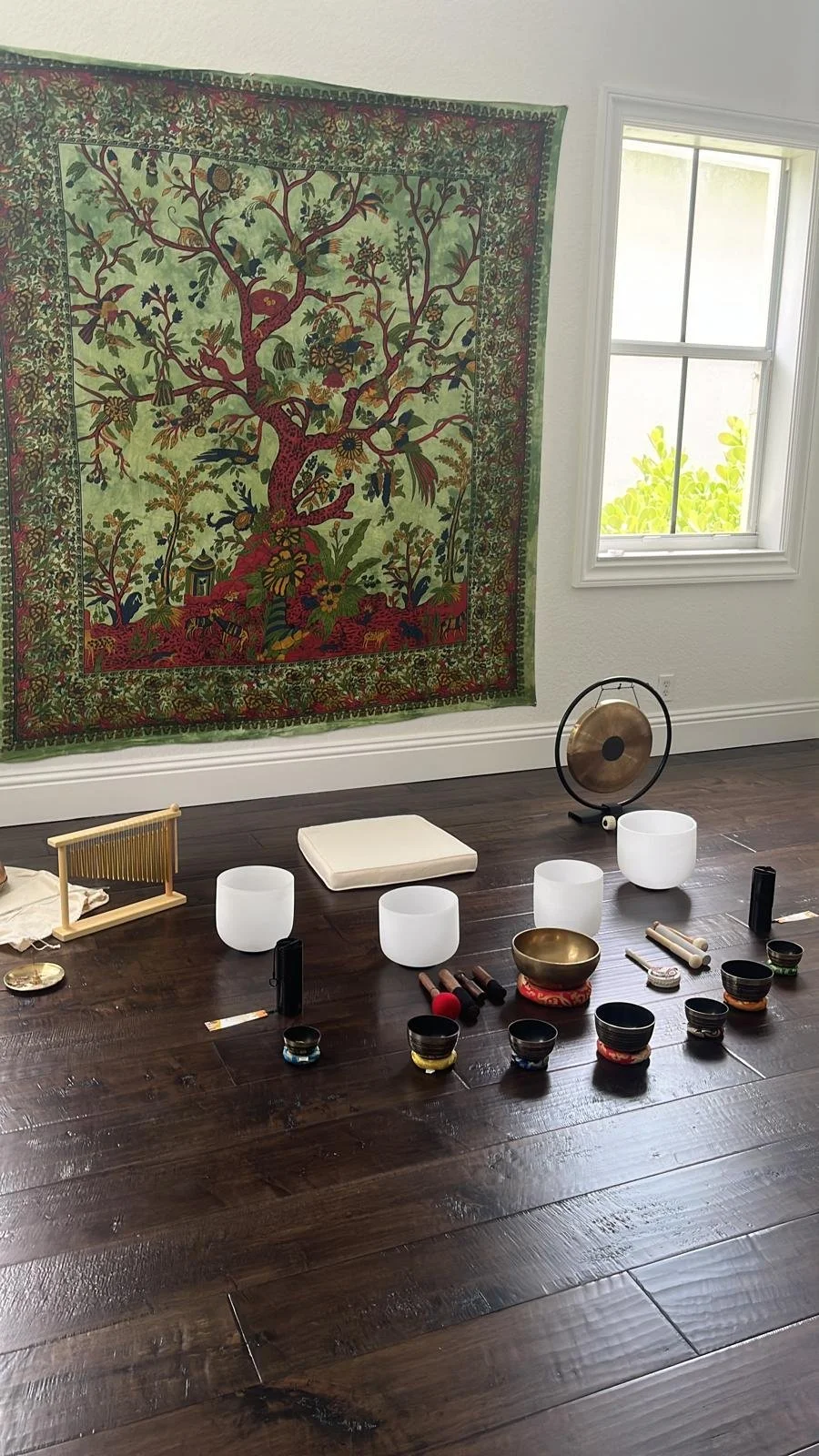 A room with a vibrant, colorful tapestry depicting a tree with birds and flowers on a wall, a window with sunlight outside, and a wooden floor with various singing bowls, a gong, and meditation accessories arranged on the floor.