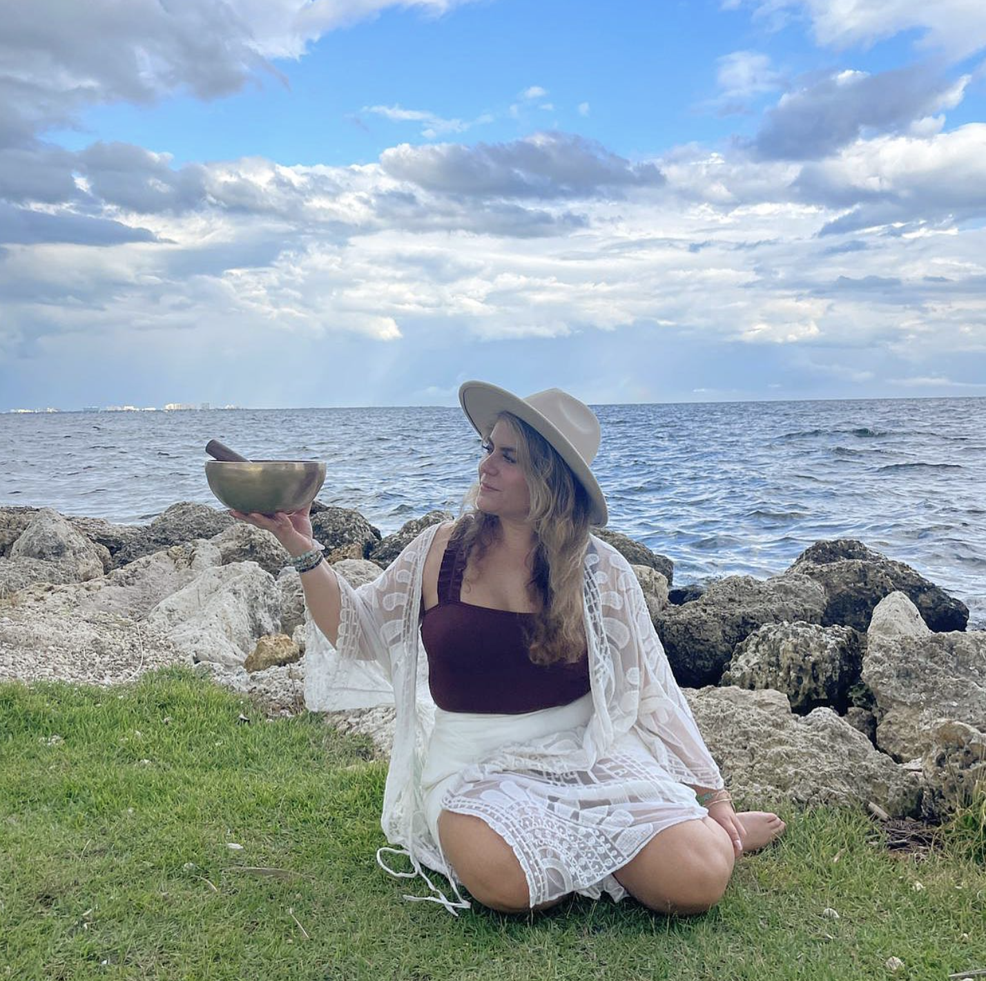 Woman sitting on grass by rocks, holding a singing bowl, wearing a wide-brim hat, white lace cover-up, and burgundy top, with ocean and cloudy sky in background.