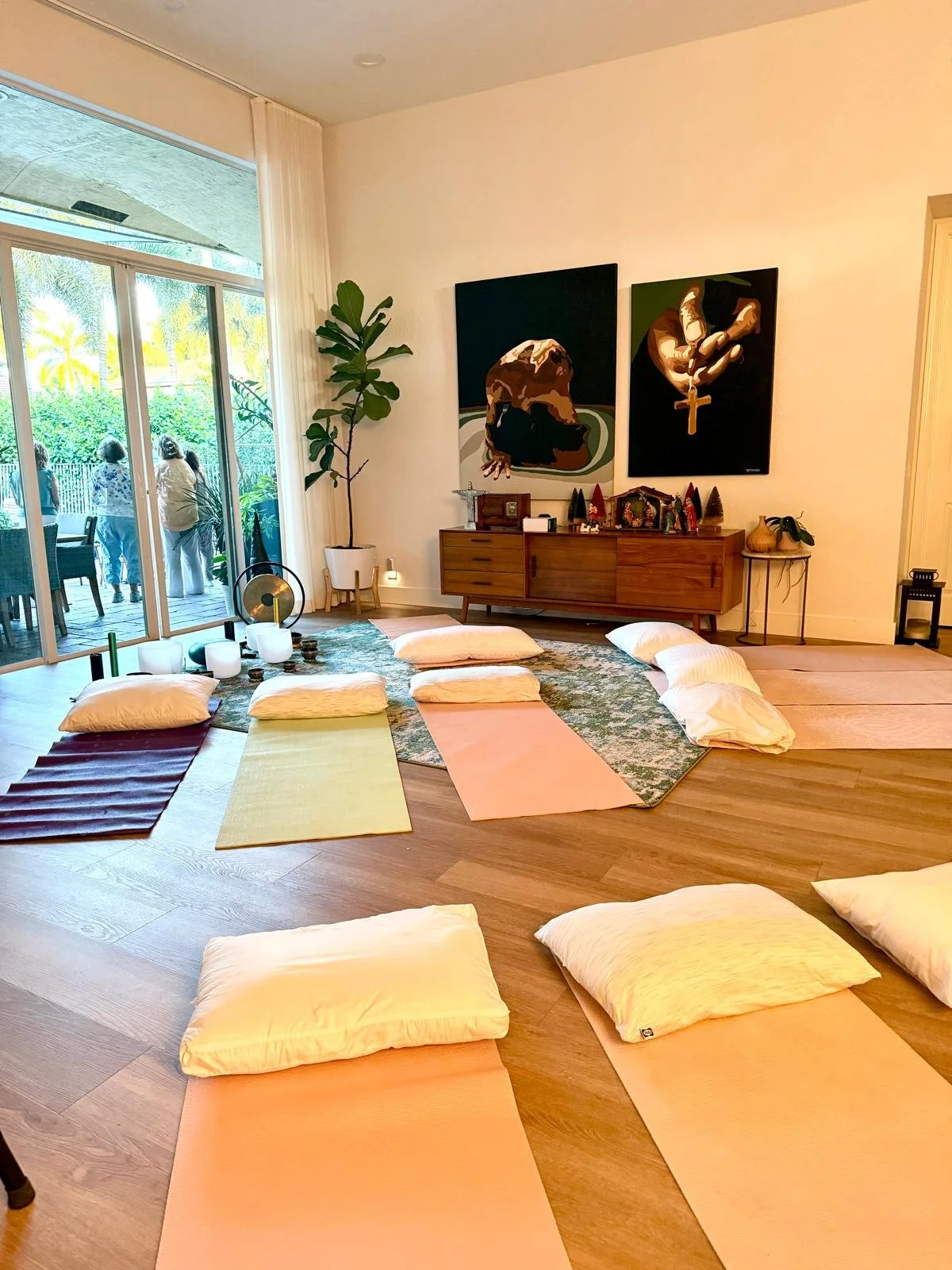 Indoor room set up for yoga with colorful mats and pillows, featuring artwork on the wall, a sliding glass door leading to a patio with people outside, and indoor plants.