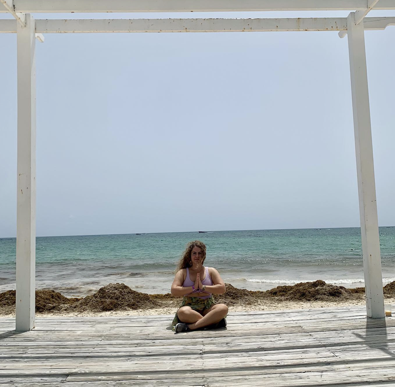 A woman sitting cross-legged on a wooden deck at the beach, with her hands in a prayer position, under a white structure, the sea and a cloudy sky in the background.