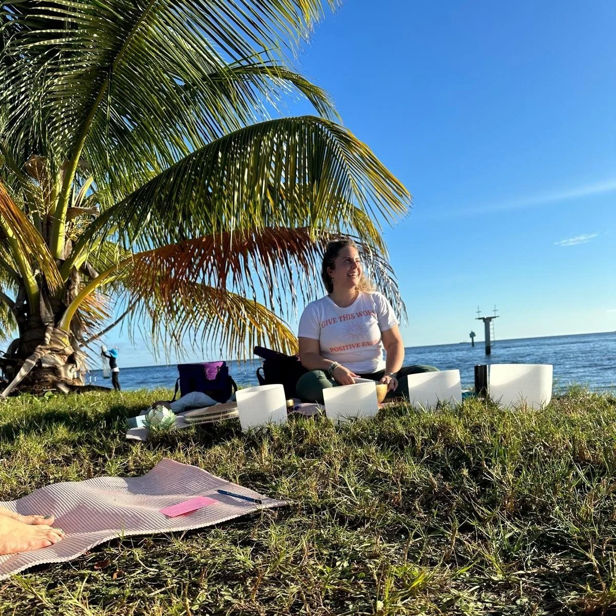 A woman sitting on the grass under a palm tree near the ocean, smiling, with singing bowls around her, during a sunny day.