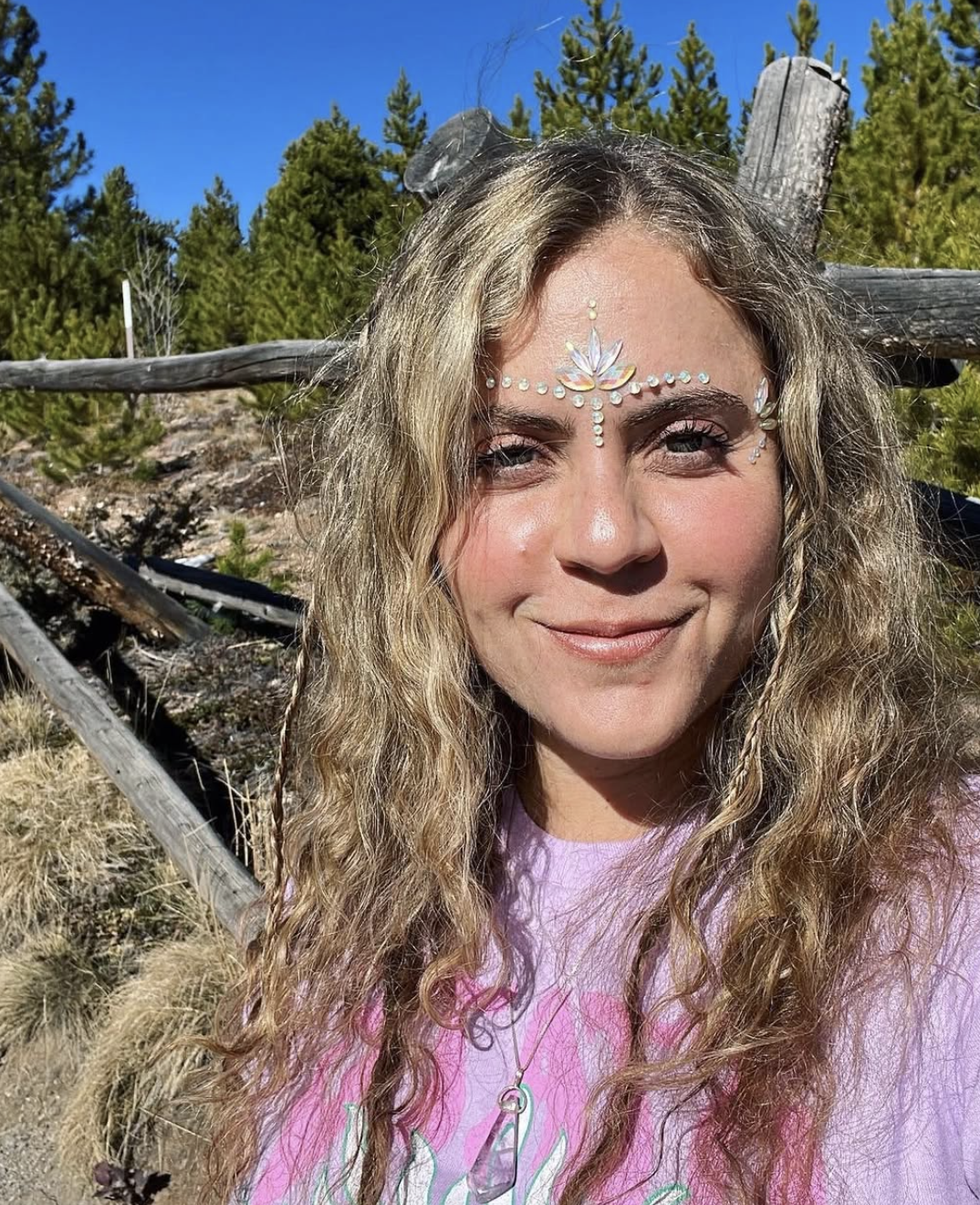 A woman with long, curly blonde hair, smiling outdoors with trees and a wooden fence in the background. She has decorative face jewelry on her forehead and wears a pink shirt.