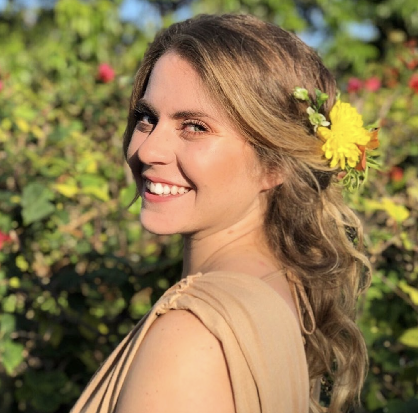 A woman with wavy hair smiling outdoors, with a yellow flower in her hair, surrounded by green foliage and flowers.
