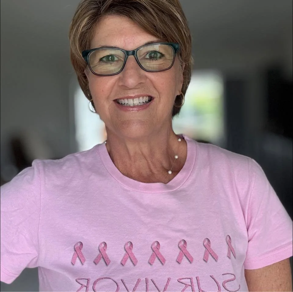 A smiling woman wearing glasses and a pink T-shirt with pink ribbons for breast cancer awareness, standing indoors with a blurred background.