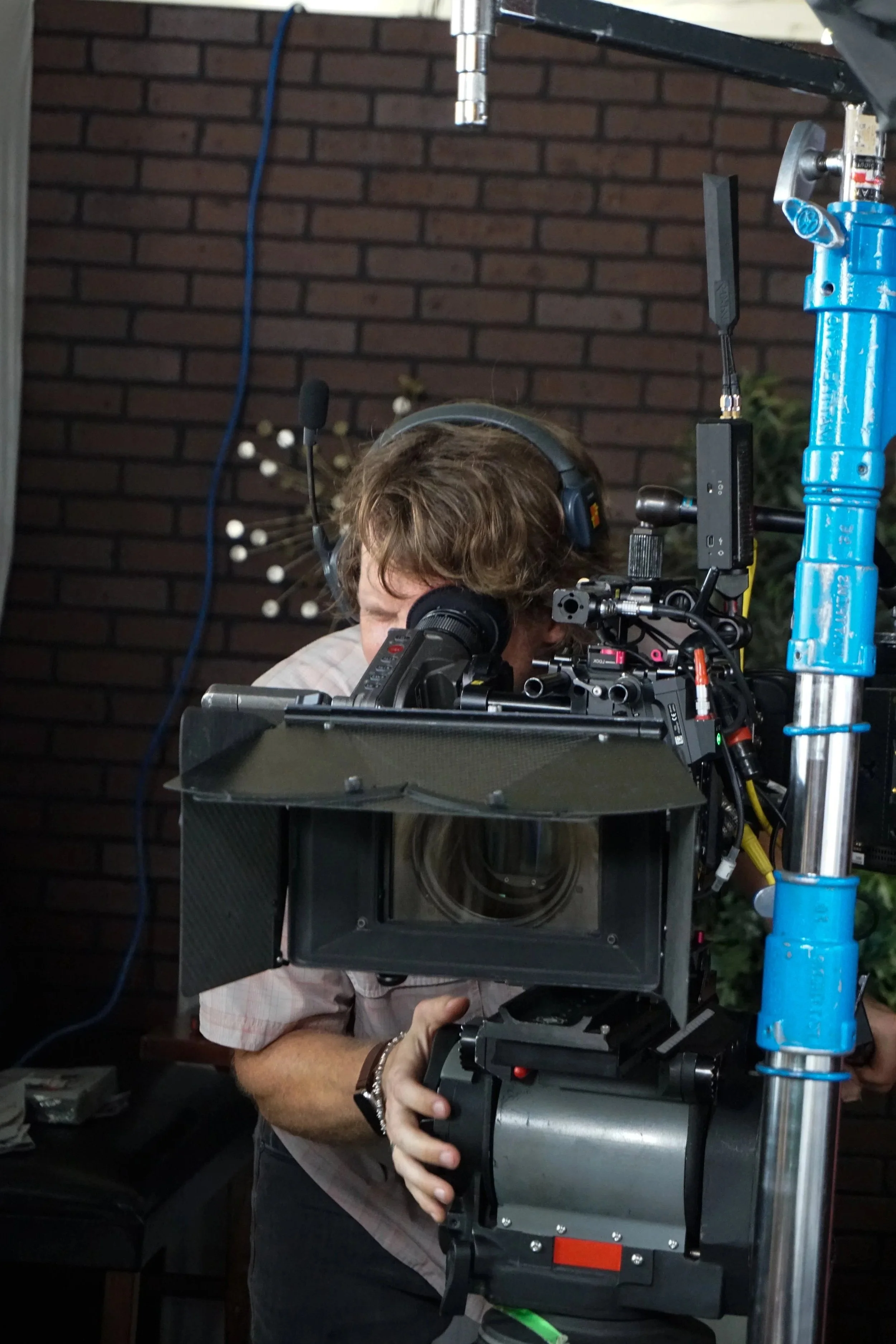 Person operating a professional film camera on a studio set with a brick wall background and green plants.